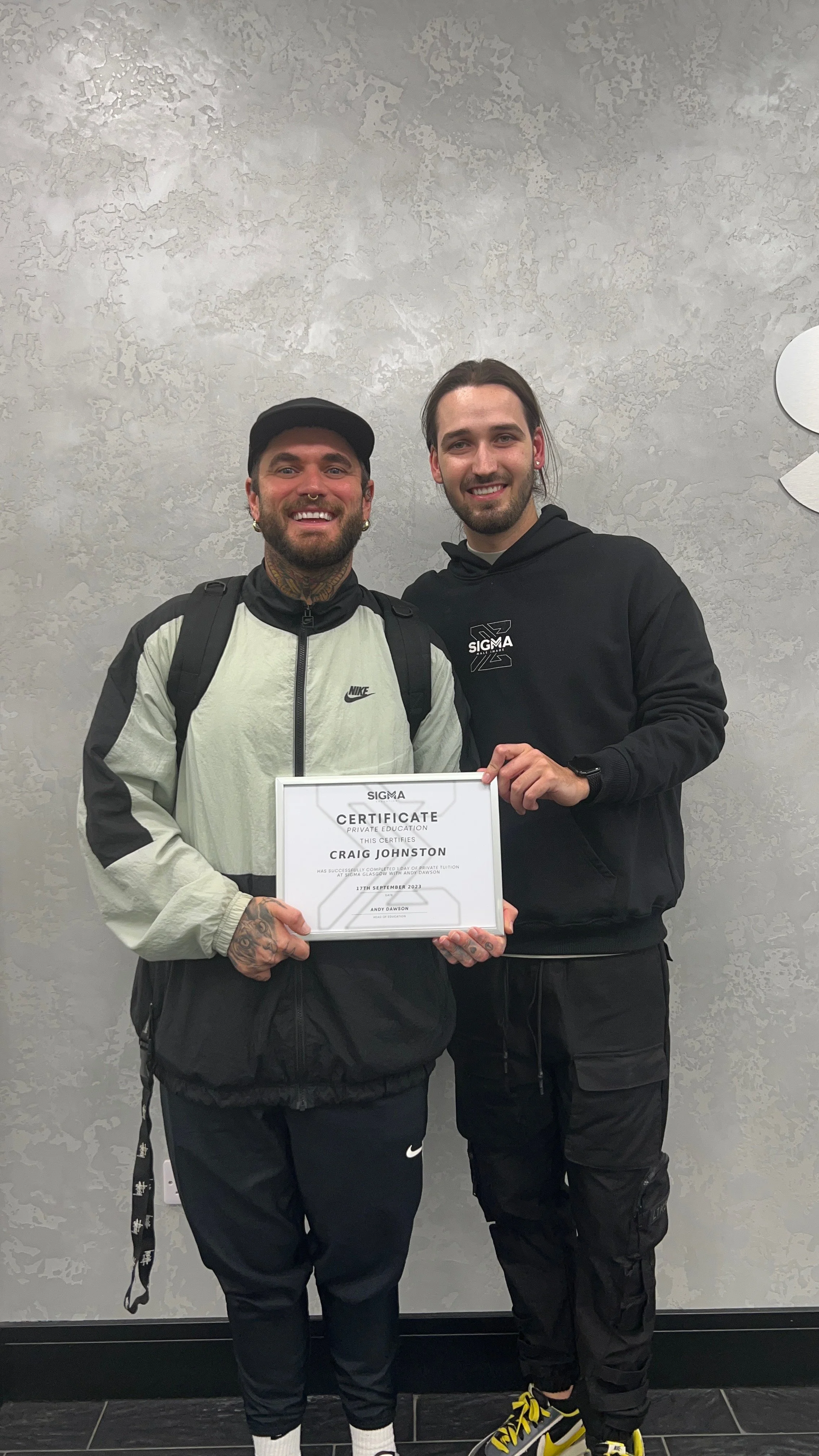 Two smiling young men standing side by side, one holding a diploma, in a gray textured wall background.