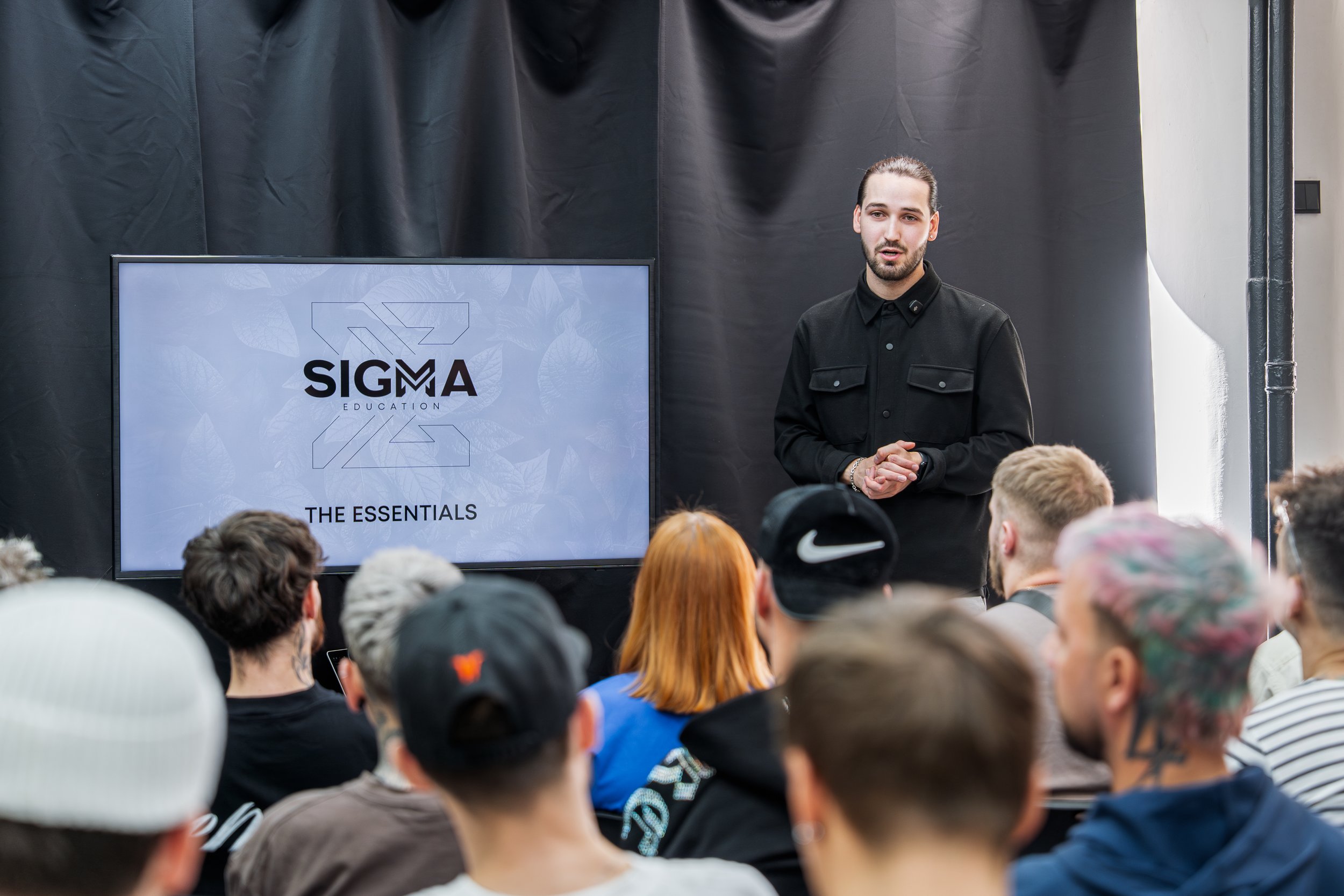 A man giving a presentation to a group of students in front of a large screen displaying 'SIGMA Education - The Essentials' recognizable logo. The audience is seated, some wearing hats and casual clothing. The setting appears to be indoors with a bla