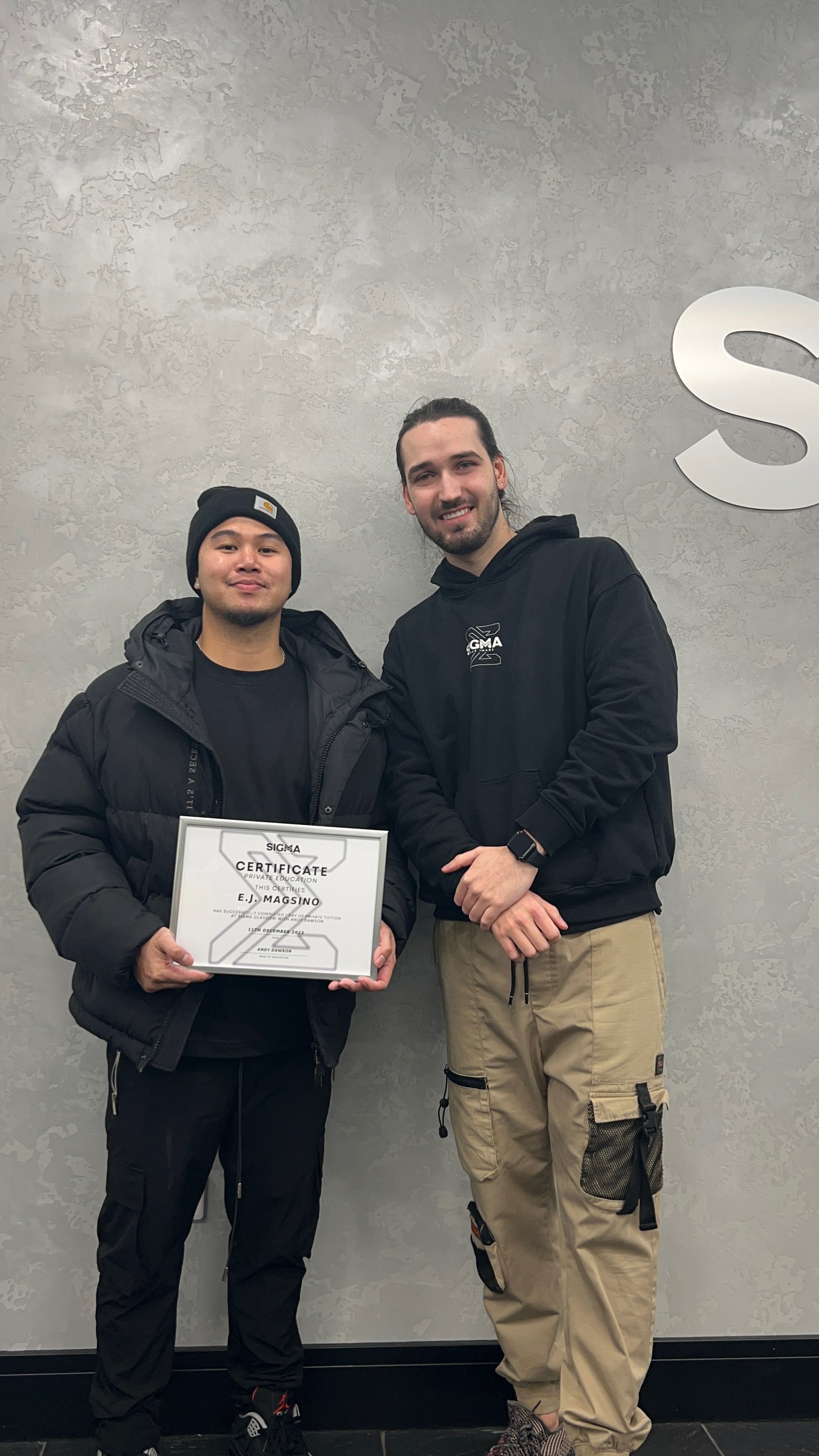 Two men standing together, one holding a certificate. They are in front of a textured gray wall with part of a large white letter S visible on the right. The man on the left is wearing a black beanie, black jacket, and black pants. The man on the rig