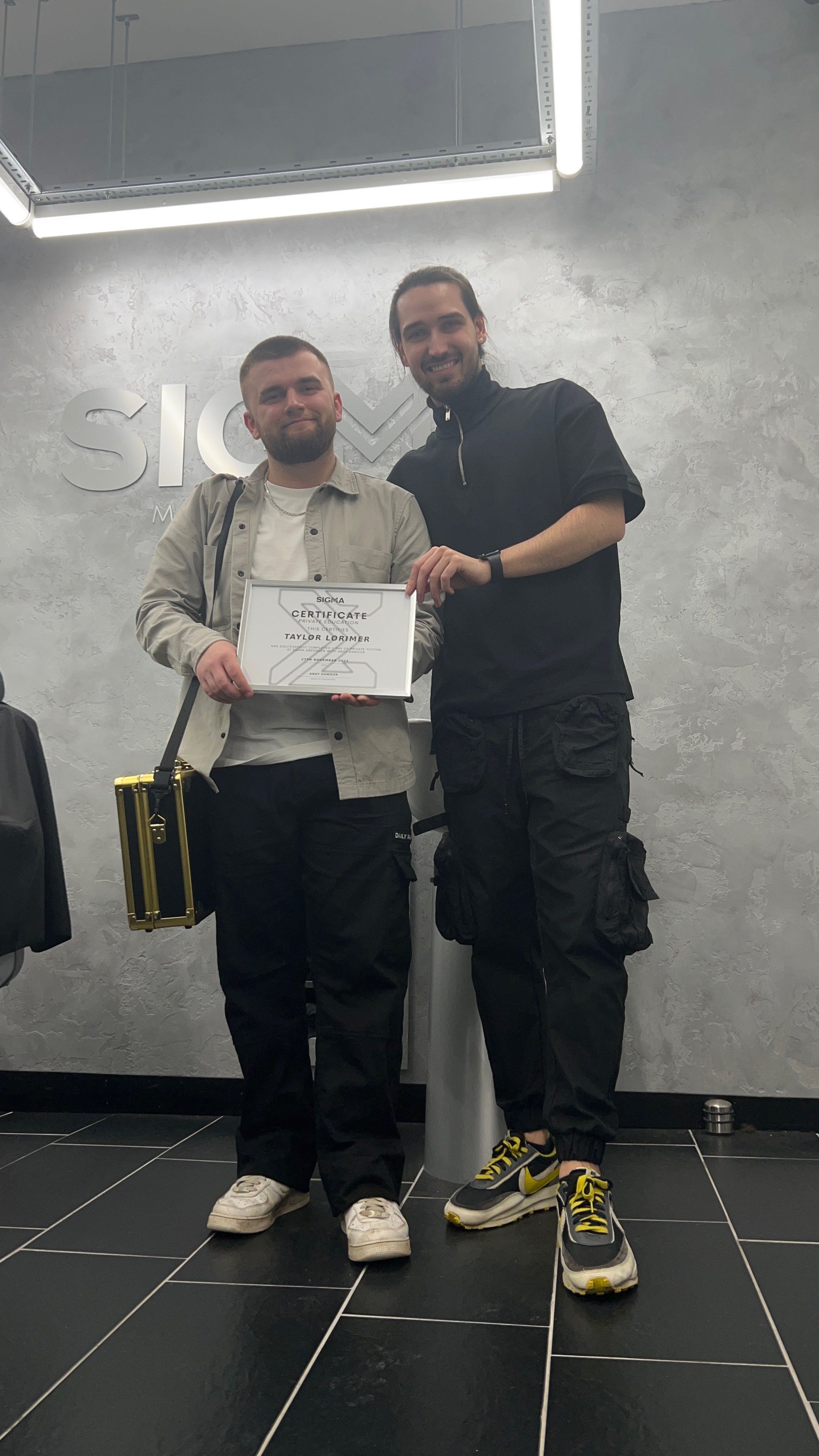 Two young men standing indoors; one holding a certificate of completion and smiling, the other smiling as well. They are in front of a wall with the word "Sigma" visible and hanging ceiling lights.