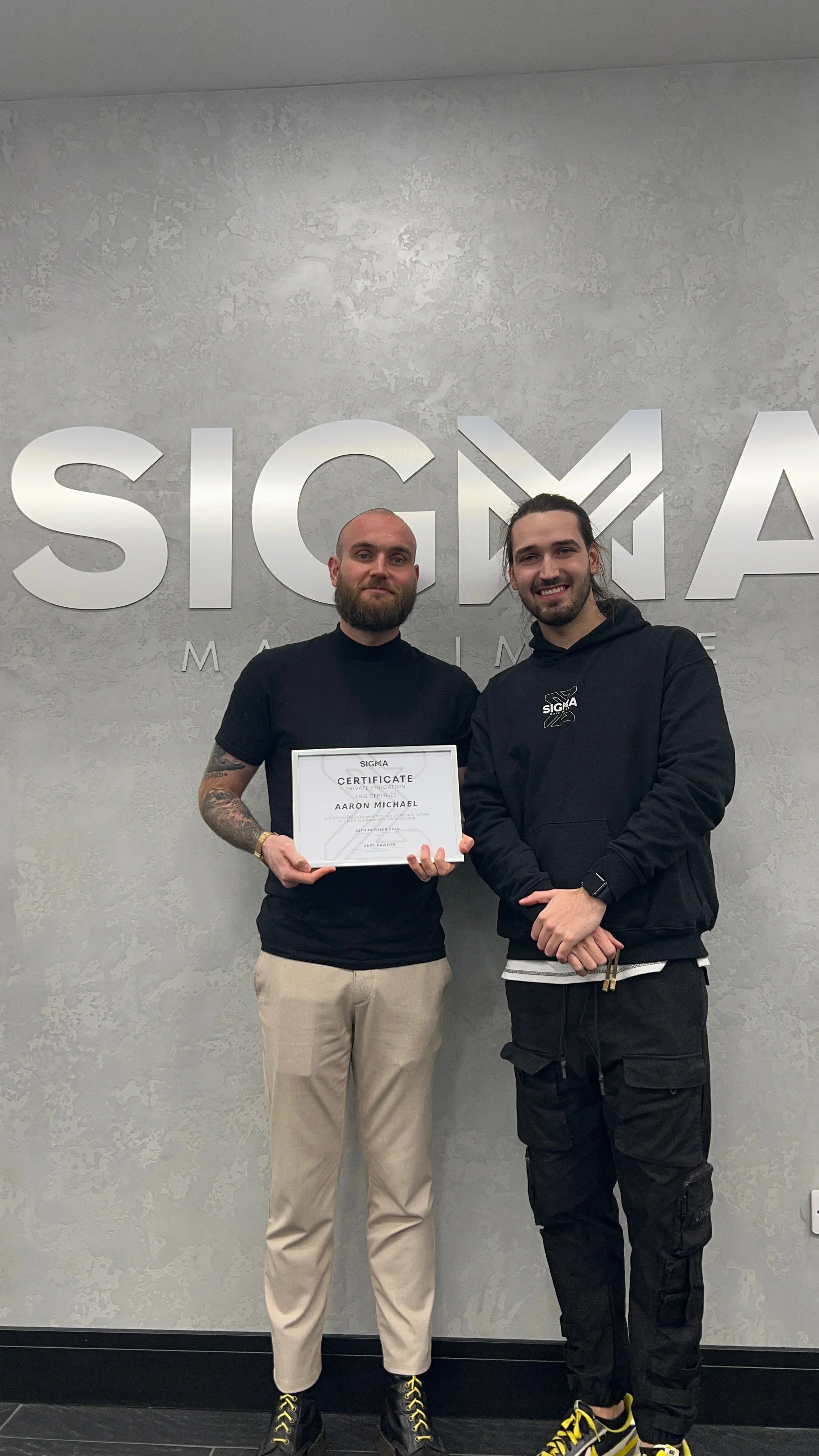 Two men standing in front of a SIGMA company sign; one is holding a certificate. Both are smiling, and the background is a textured gray wall.