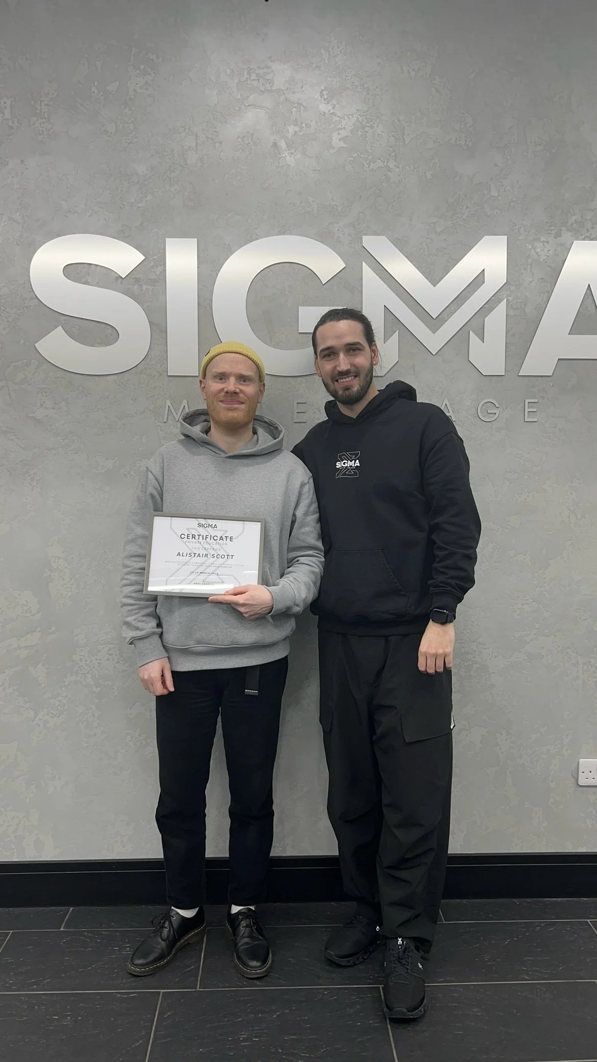 Two men standing in front of a wall with the SIGMA logo; the man on the left is holding a certificate, and both are smiling at the camera.