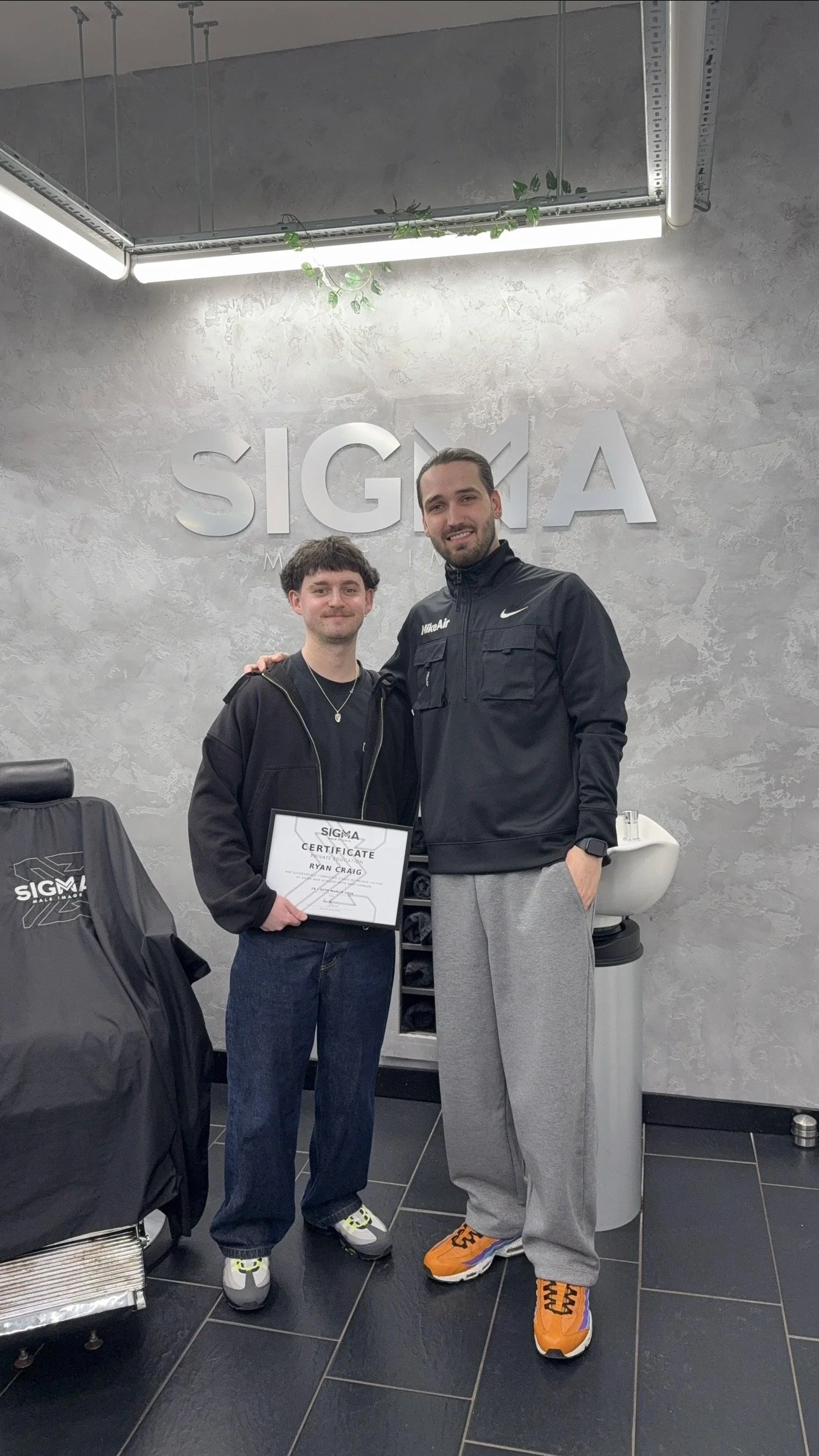 Two men standing inside a room, one of whom is holding a certificate. The background features a large sign that reads 'SIGMA'. The setting appears to be a professional or celebration event, with modern decor and lighting.