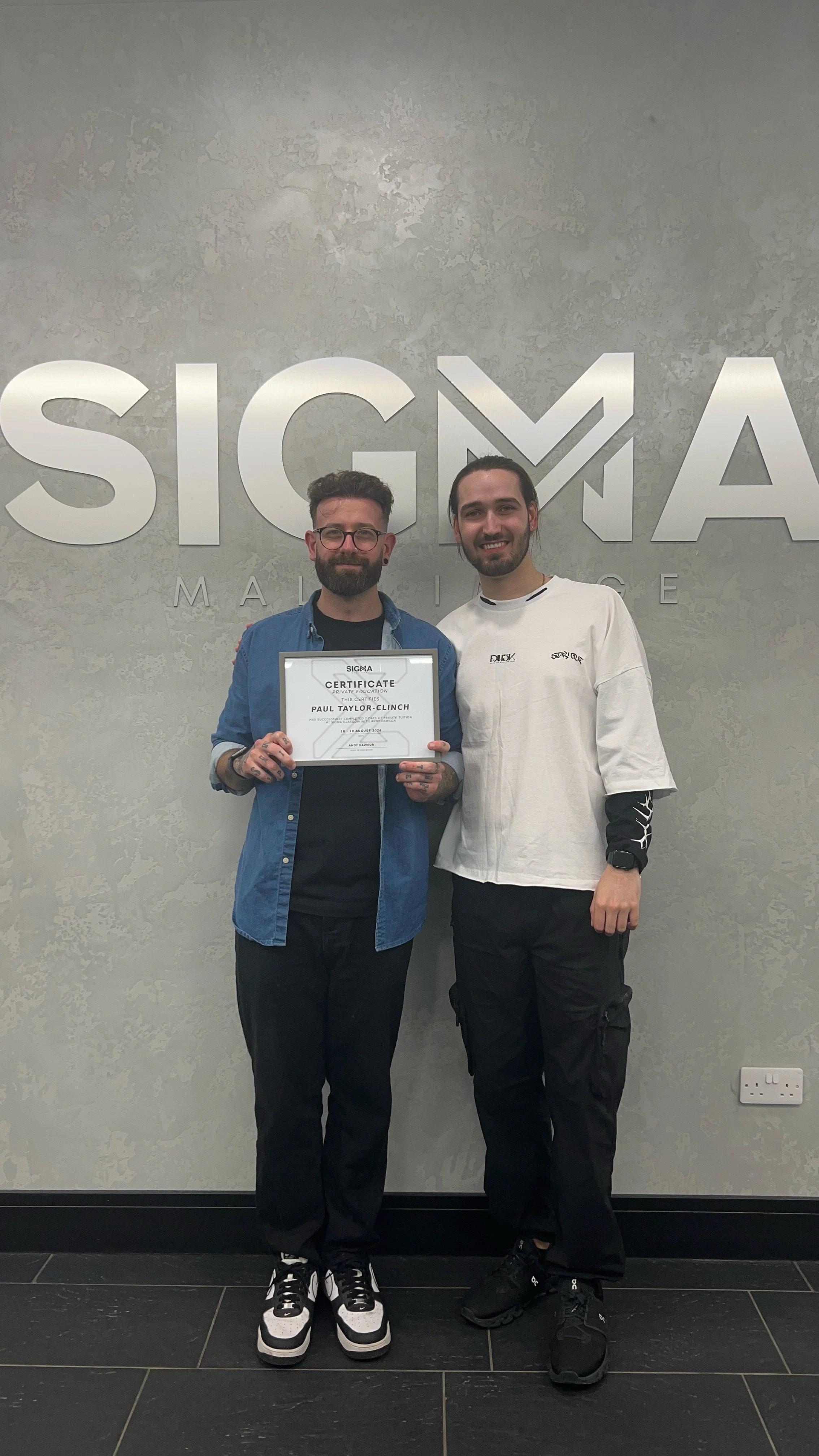 Two young men standing in front of a wall with the Sigma logo. One man is holding a certificate.