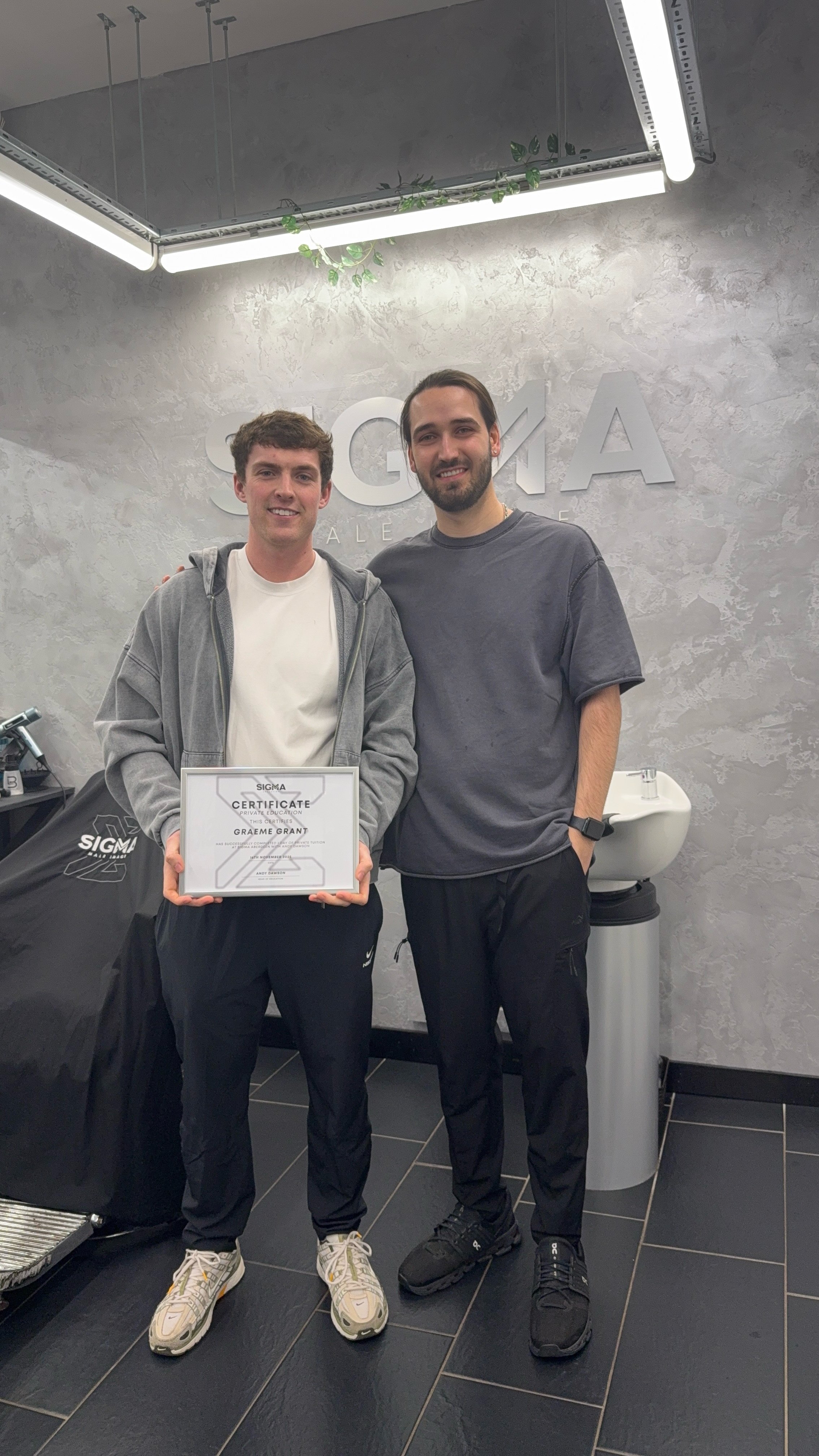 Two young men standing in a modern, gray-walled room, smiling at the camera. The man on the left is holding a framed certificate. The background shows a wall with the word 'SIGMA' and a sink.