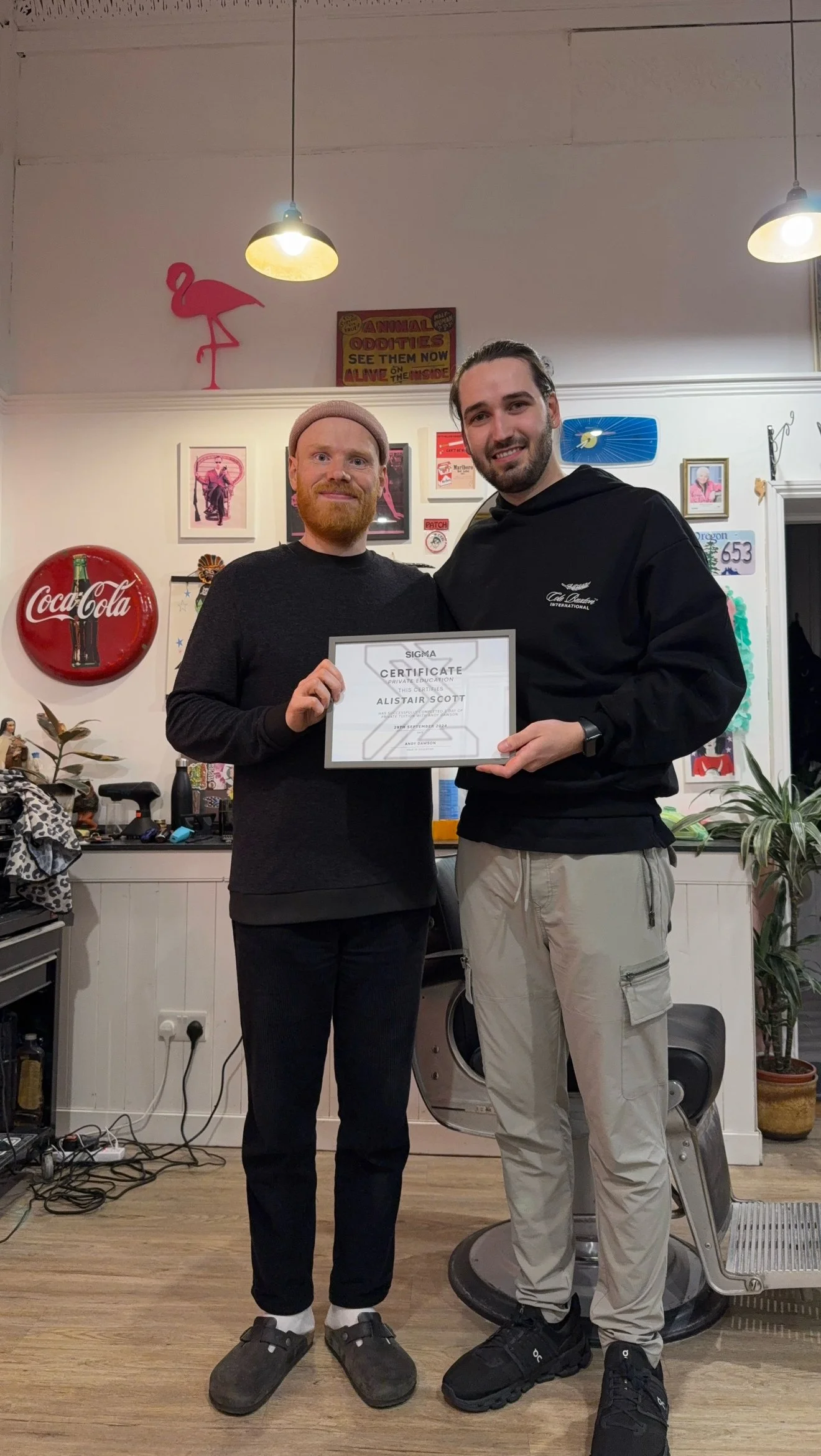Two men standing inside a barbershop, holding a certificate. The man on the left has a beard and is wearing a beanie, black shirt, and black pants. The man on the right has a beard and long hair tied back, wearing a black hoodie and beige cargo pants