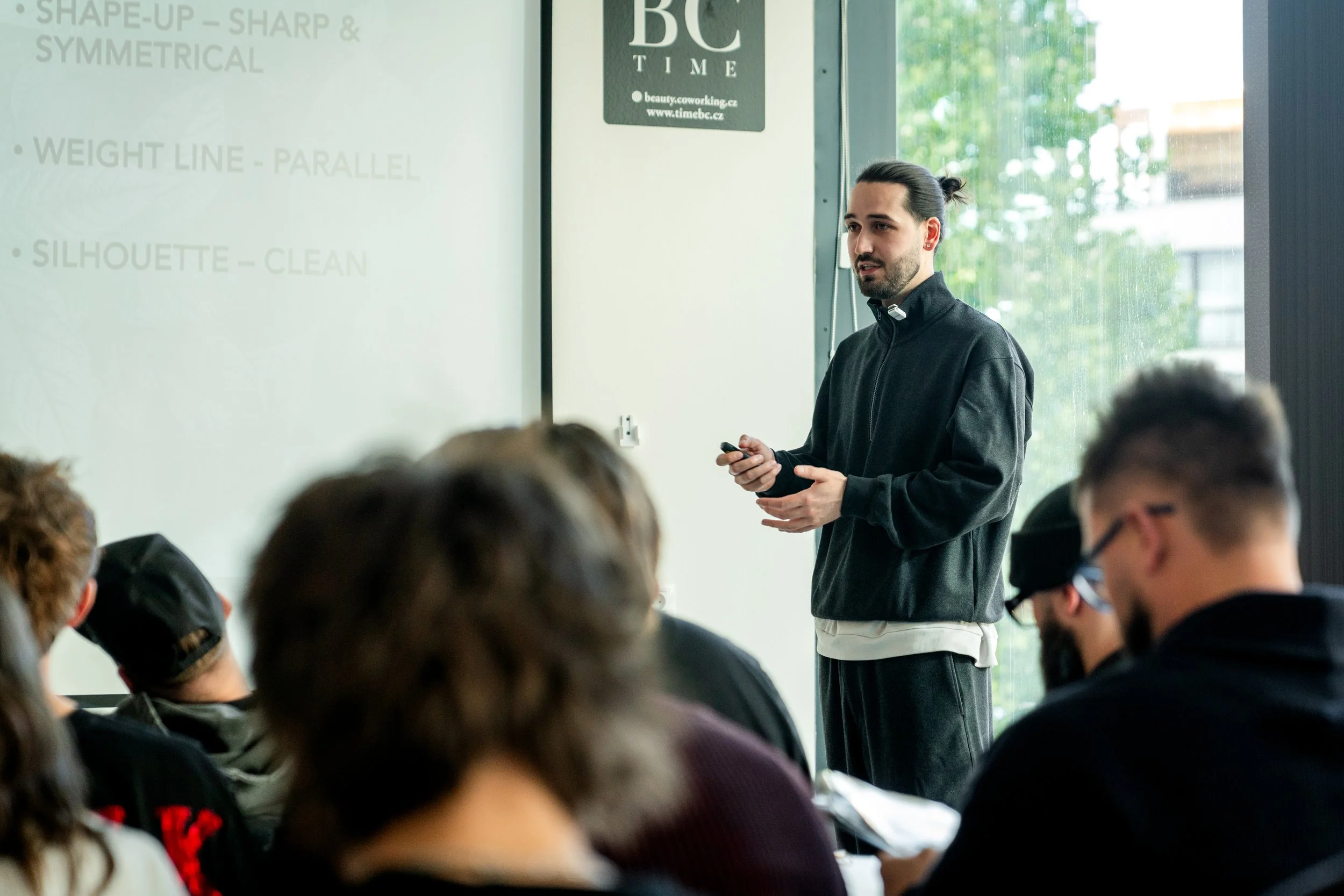 A man giving a presentation in a classroom or conference room with a whiteboard showing bullet points, and several audience members seated and listening.
