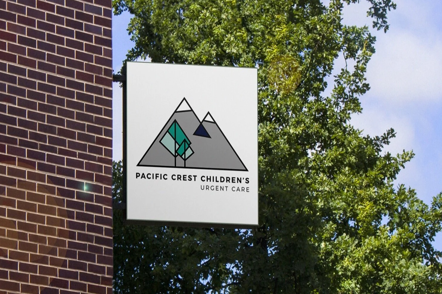 Sign for Pacific Crest Children's Urgent Care with a mountain logo and stylized trees, located outdoors near green trees and a brick building.