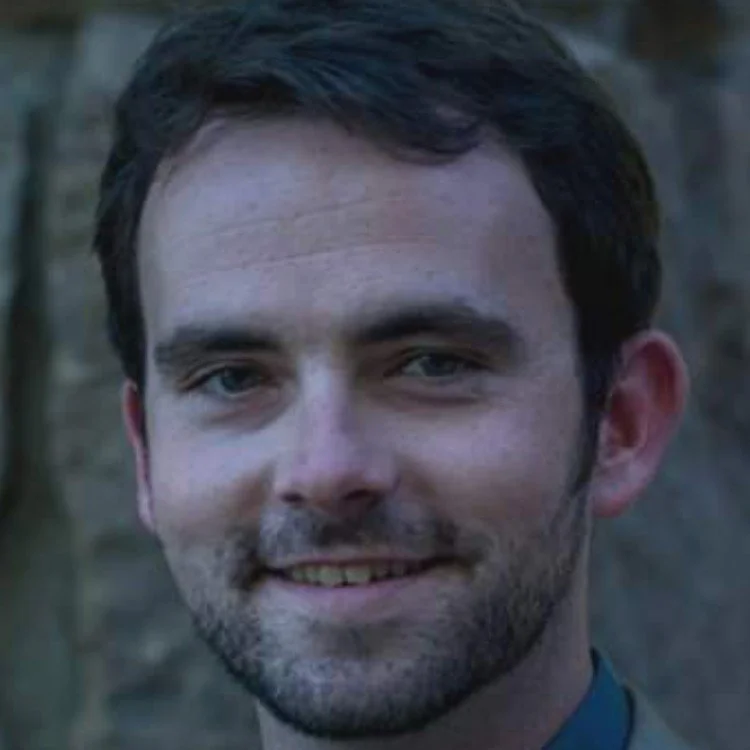 Close-up of a man with dark hair and a beard smiling outdoors.