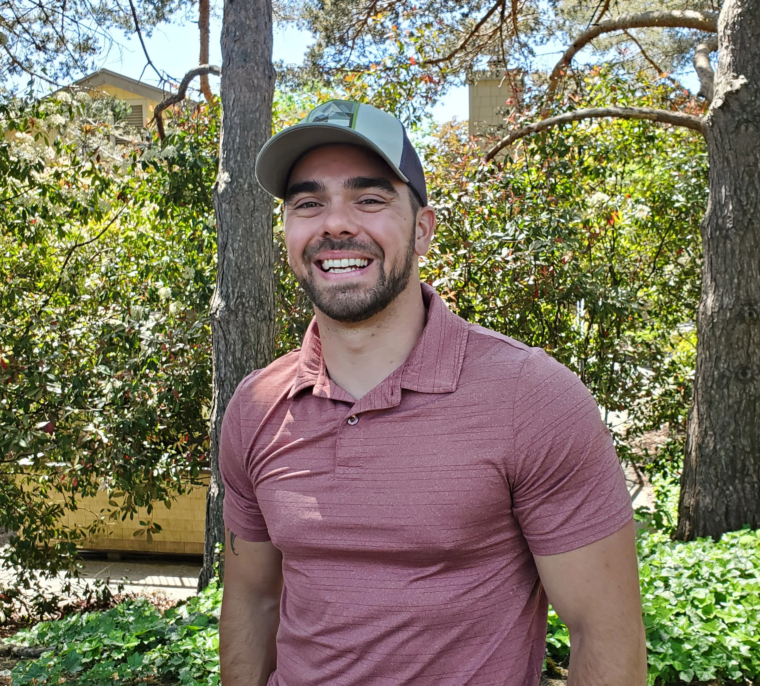 A smiling man wearing a baseball cap and a maroon shirt standing outdoors among trees and bushes.