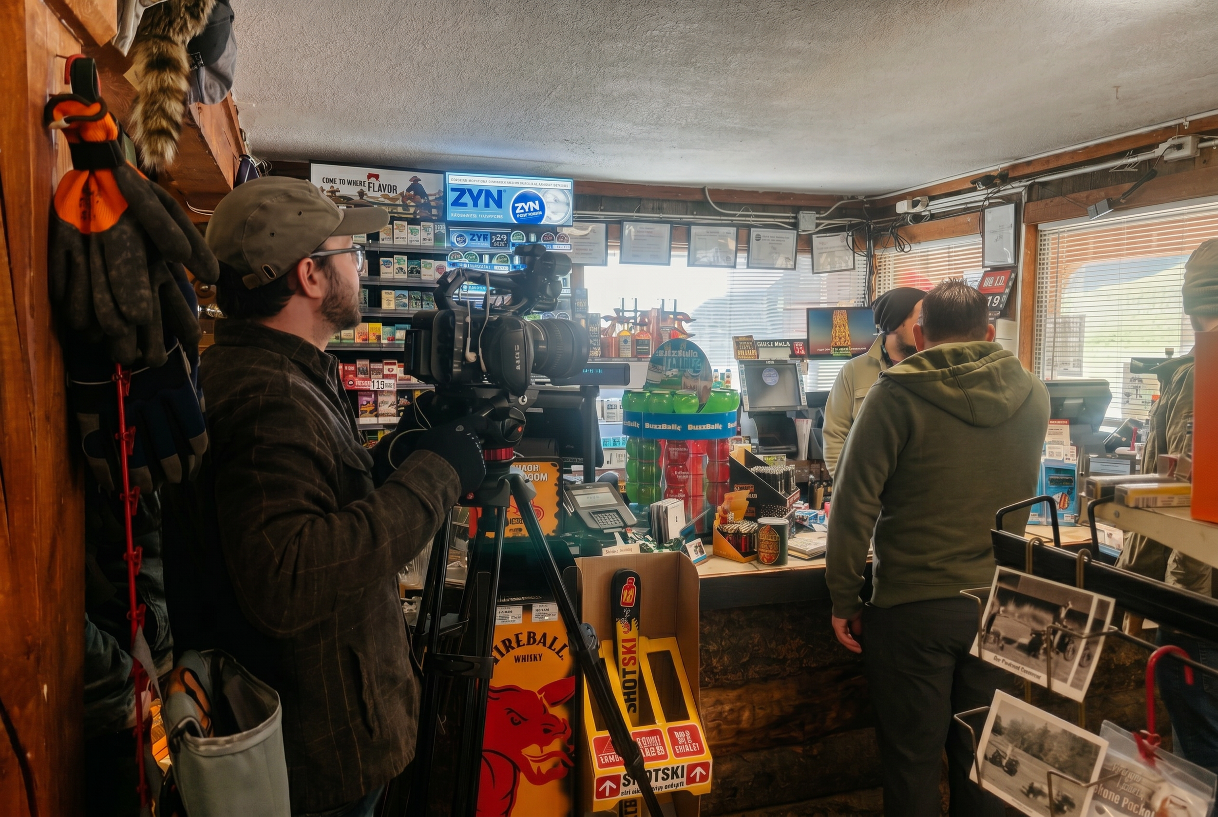 Inside a retail store with people, shelves, and a camera crew filming. There are signs, displays, and assorted items on the shelves and counters.