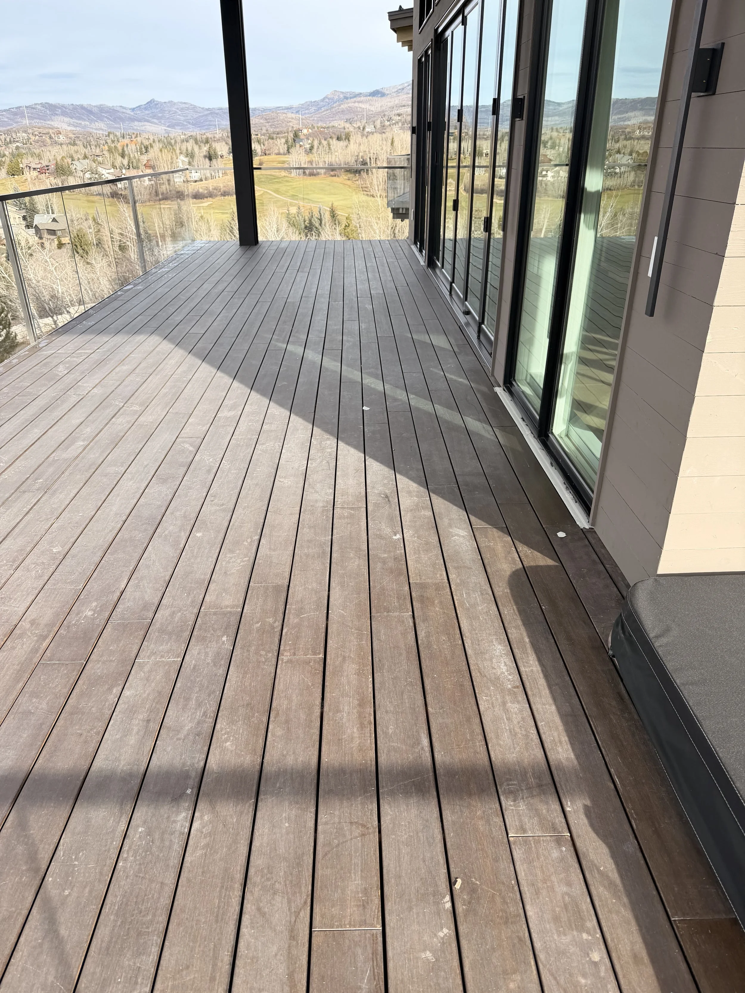 A spacious outdoor balcony with wooden plank flooring and glass railing, overlooking a scenic landscape of rolling hills, trees, and distant mountains under a cloudy sky.