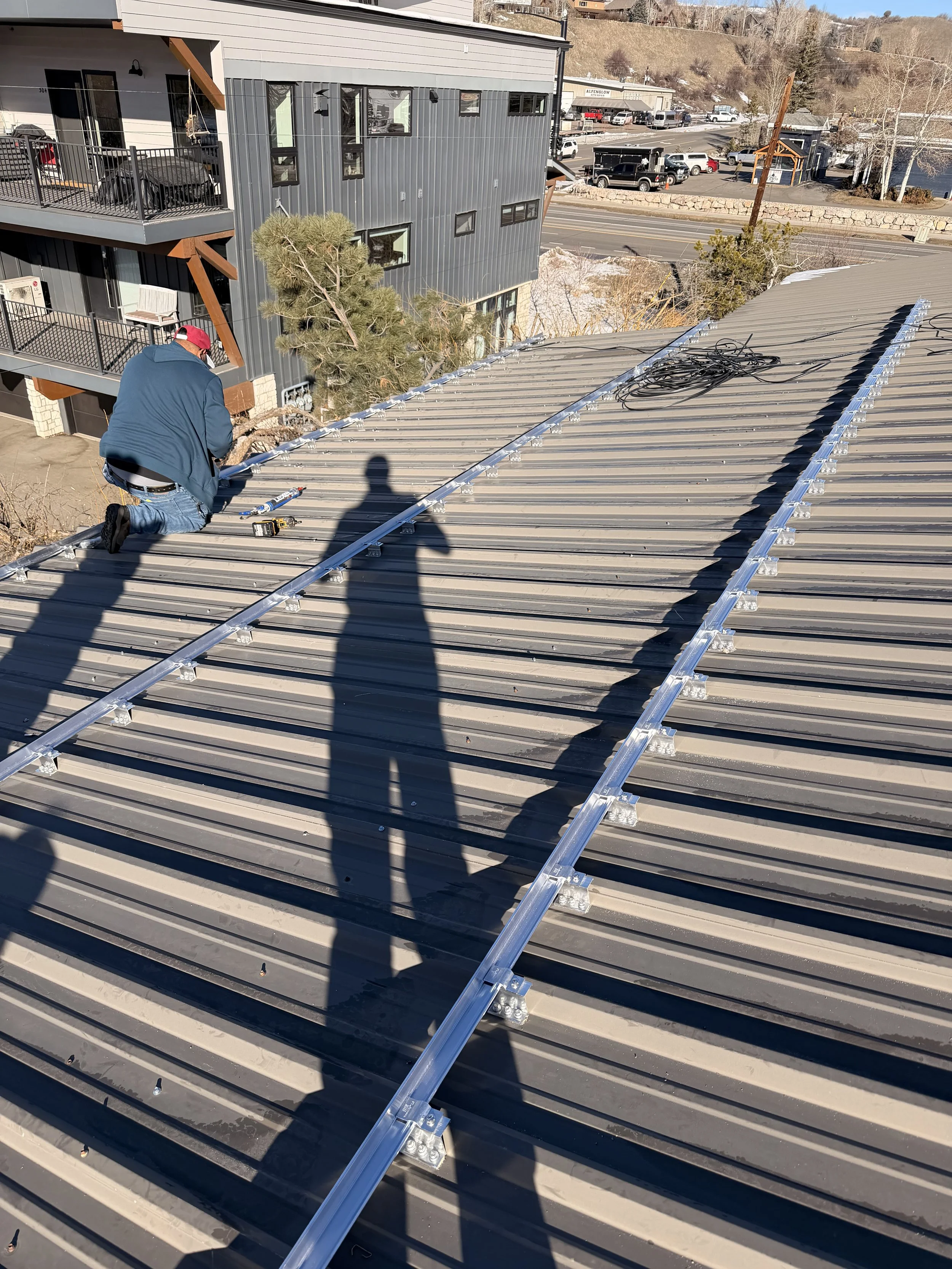 A person kneeling on a metal roof installing or repairing a solar panel mounting system, with tools scattered nearby and a shadow of another person taking the photo.