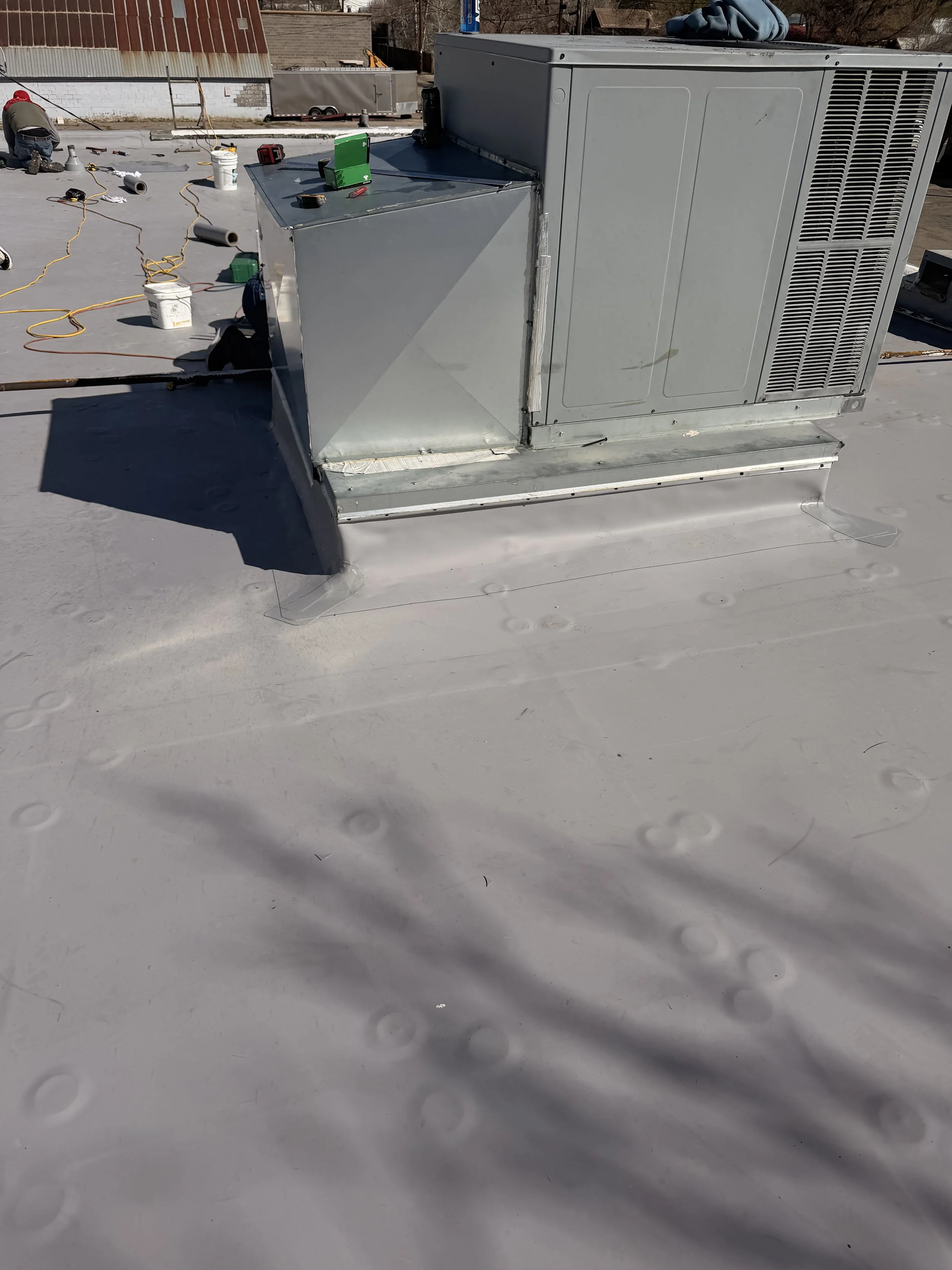 A rooftop with air conditioning units under construction, showing tools, wires, and equipment scattered around, with a person working in the background.
