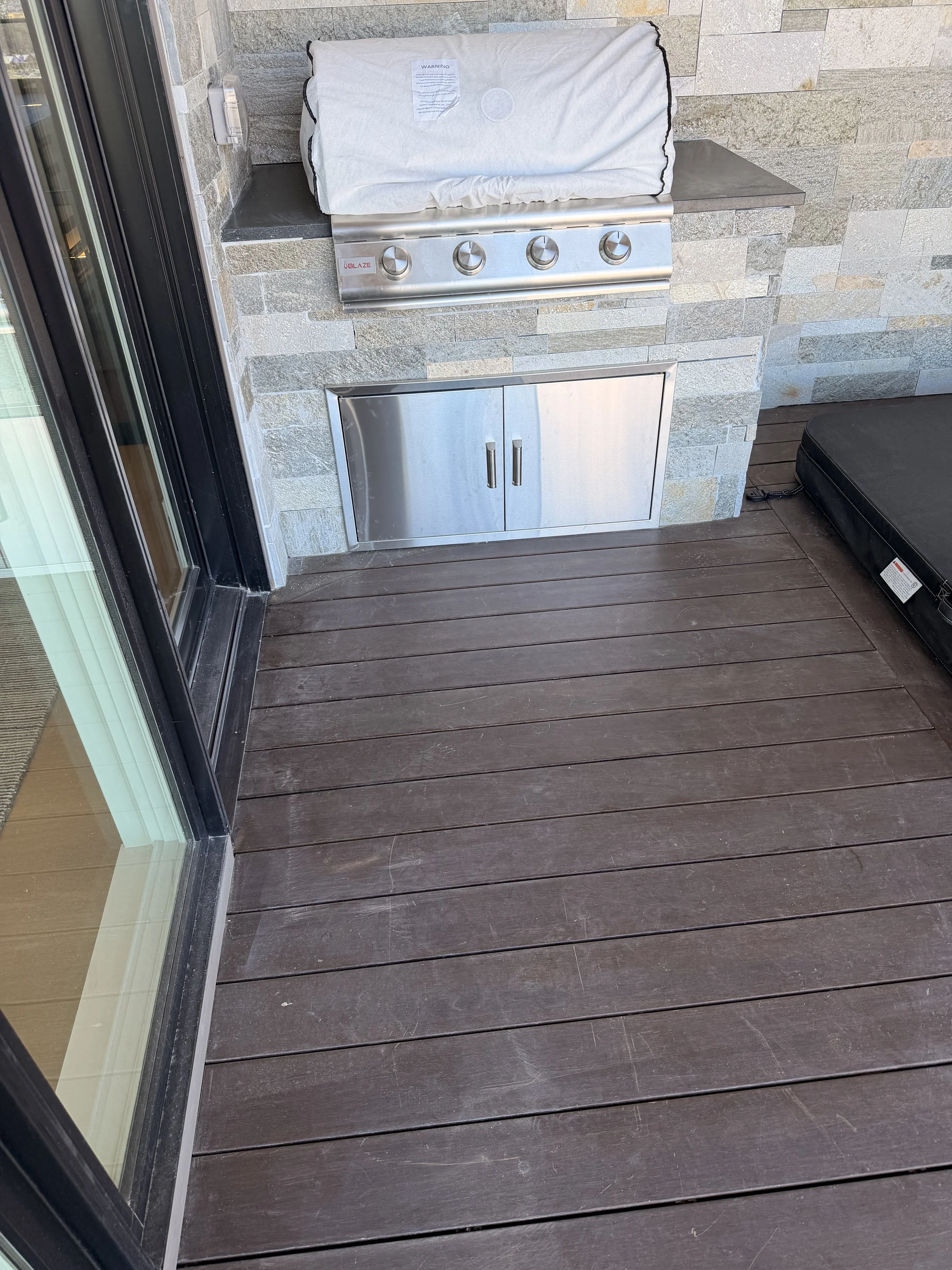 An outdoor balcony with a wooden floor, a stone wall, a stainless steel barbecue grill covered with a white cloth, a small stainless steel storage cabinet below, and a black cushioned chair on the right.
