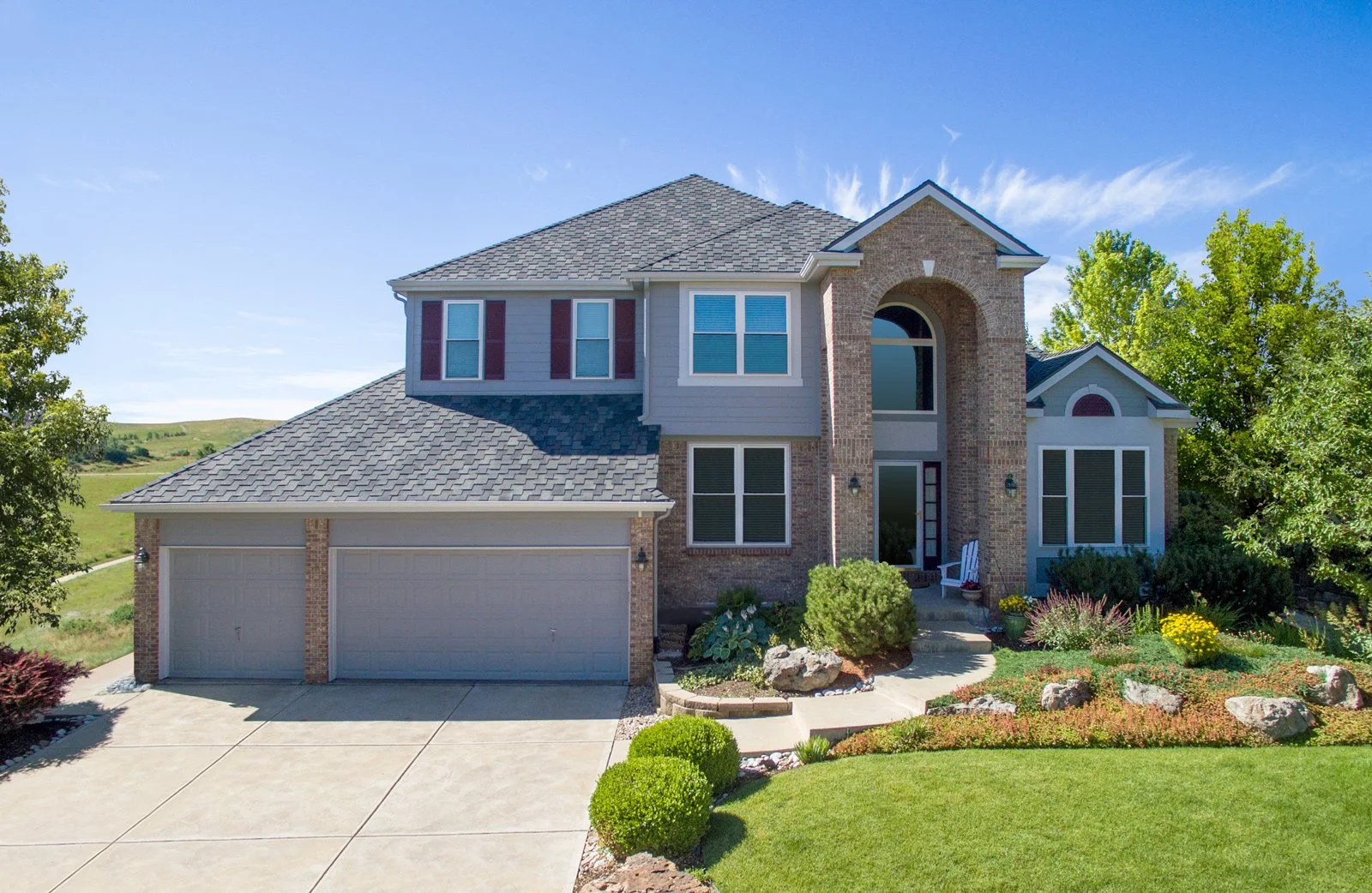 A two-story house with a brick and gray siding exterior, a three-car garage, landscaped front yard, and blue sky with scattered clouds.