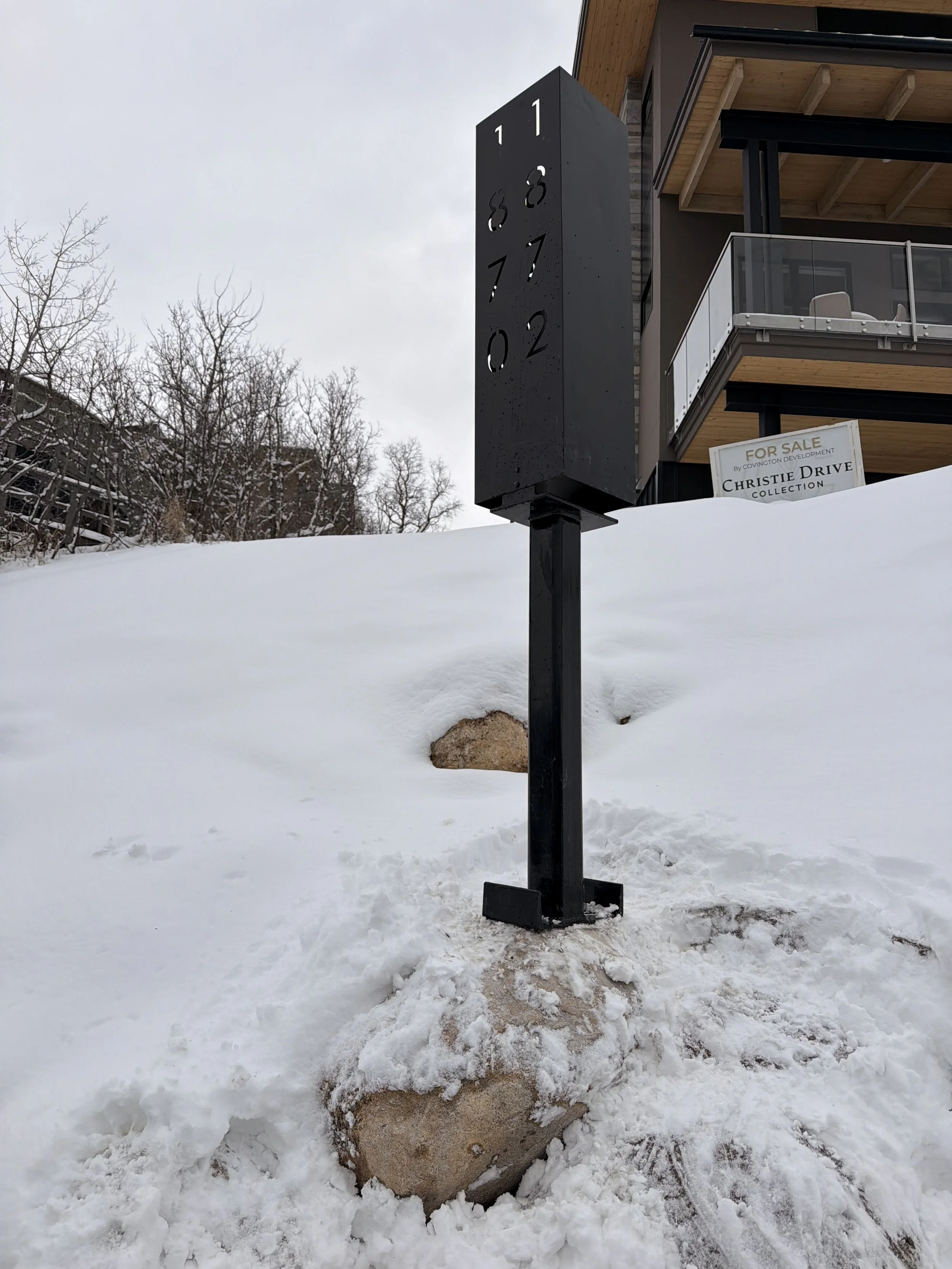 A black post with cutout numbers 0 to 11 resembling a clock face stands in the snow in front of a modern building with balconies. A 