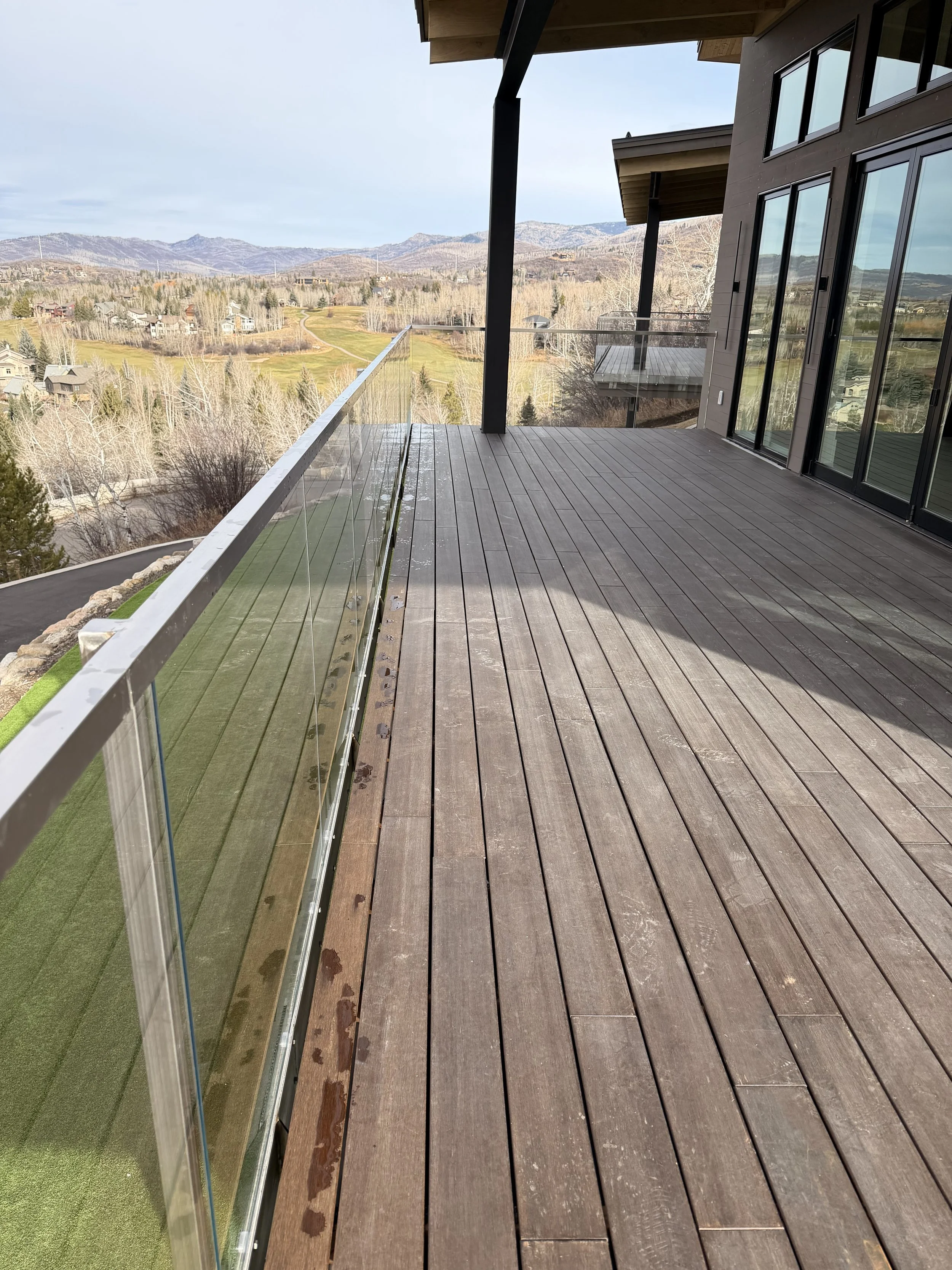 View from a balcony with glass railing, wooden floor, and sliding glass doors, overlooking a scenic landscape with trees, hills, and distant mountains under a cloudy sky.