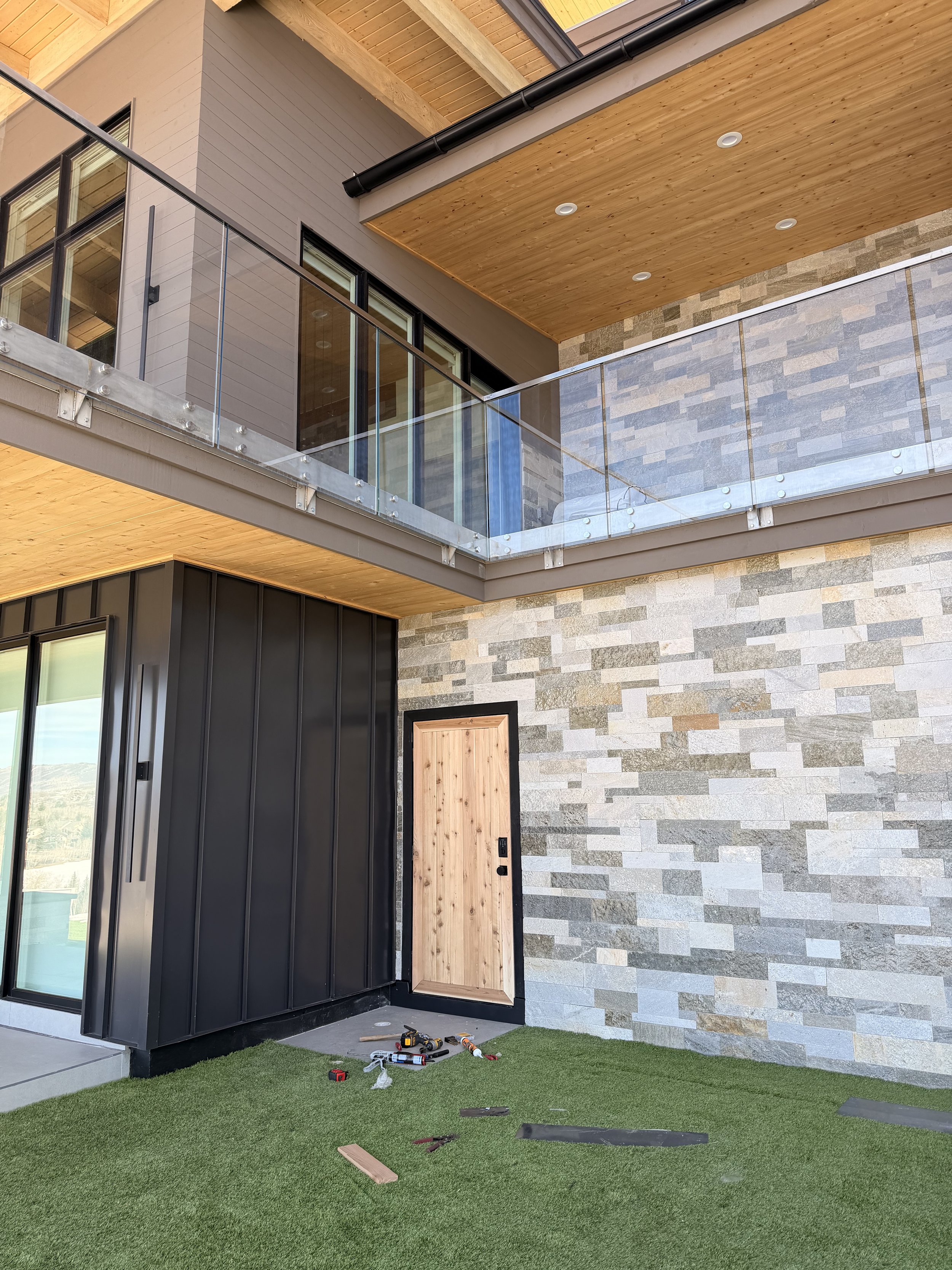 Modern house exterior with stone wall, black metal siding, wooden door, glass railing, and artificial grass lawn, with tools and materials on the ground indicating ongoing construction or renovation.