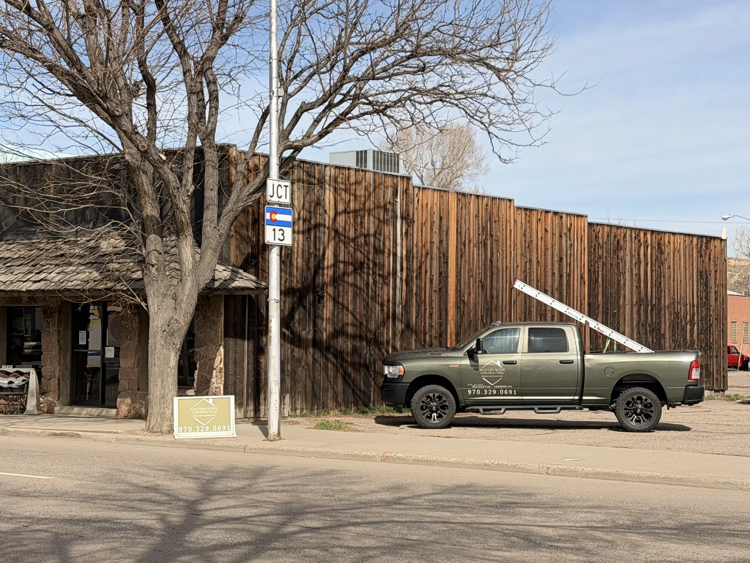 Street view with a leafless tree, a wooden building, a parking lot, and a pickup truck parked in front. There is a street sign indicating a junction, and a small sign advertising a local contracting company with a phone number.