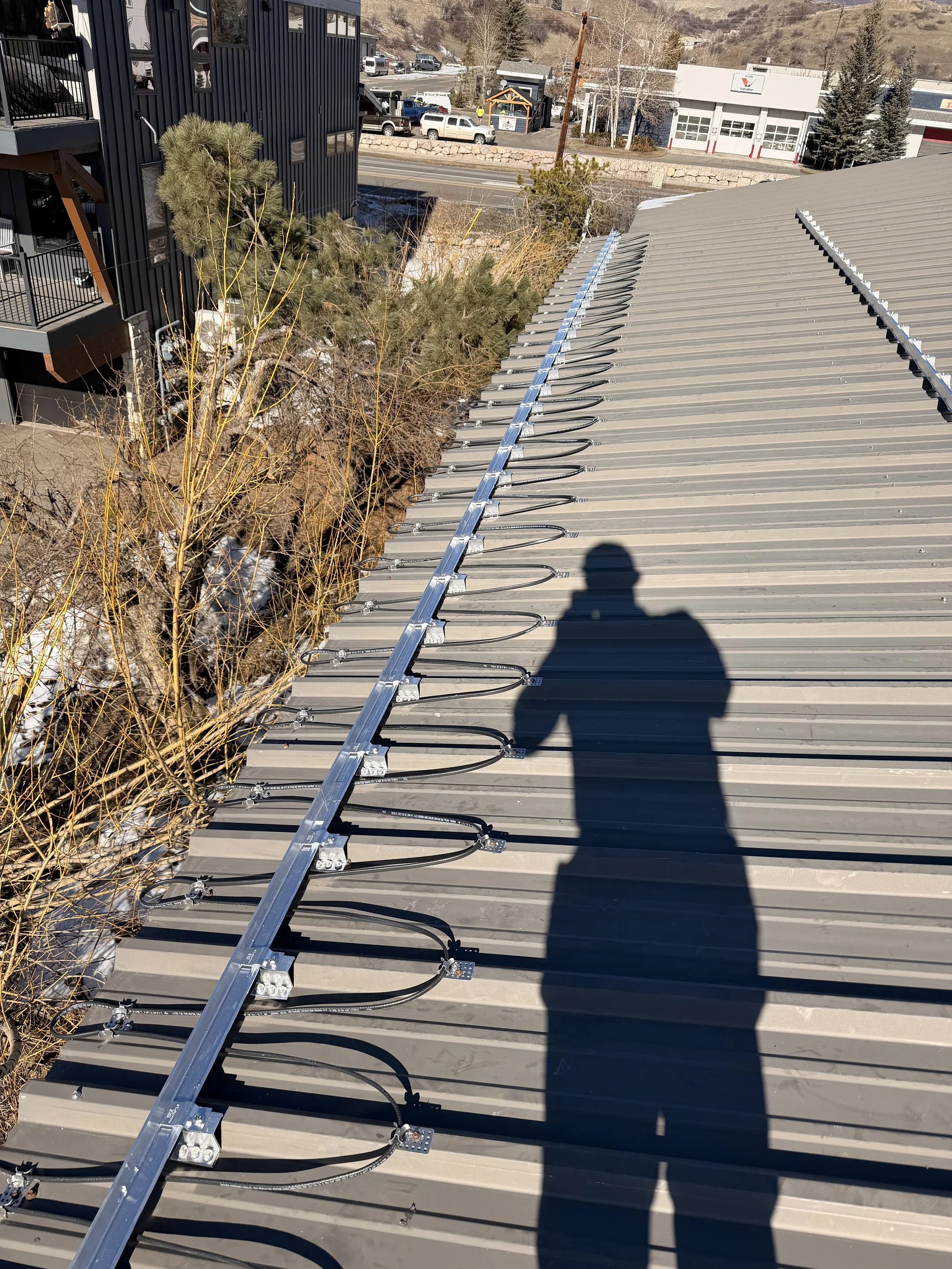 Metal roof with solar panel mounting rails and wiring, shadow of person taking the photo, nearby trees and commercial buildings in a suburban area.
