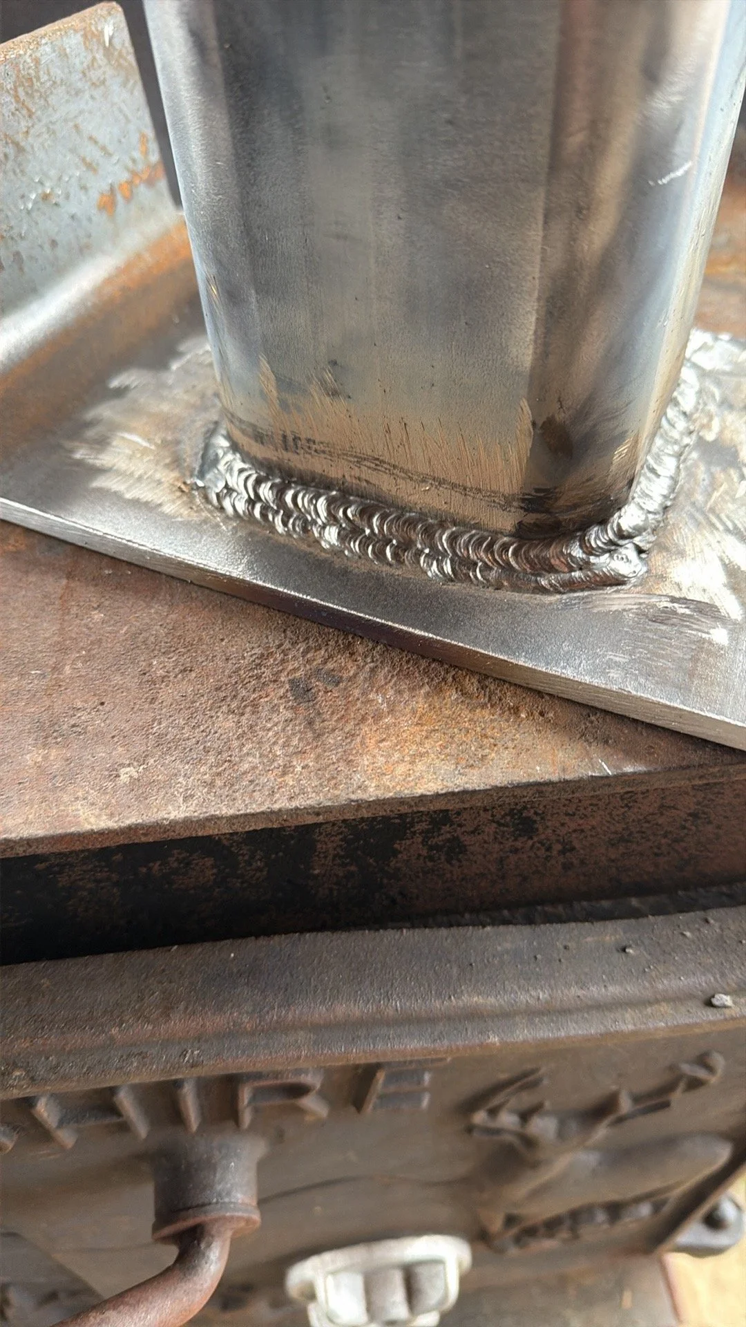 Close-up of a metal welding joint on a metal structure, with a rough, rusty surface visible nearby.