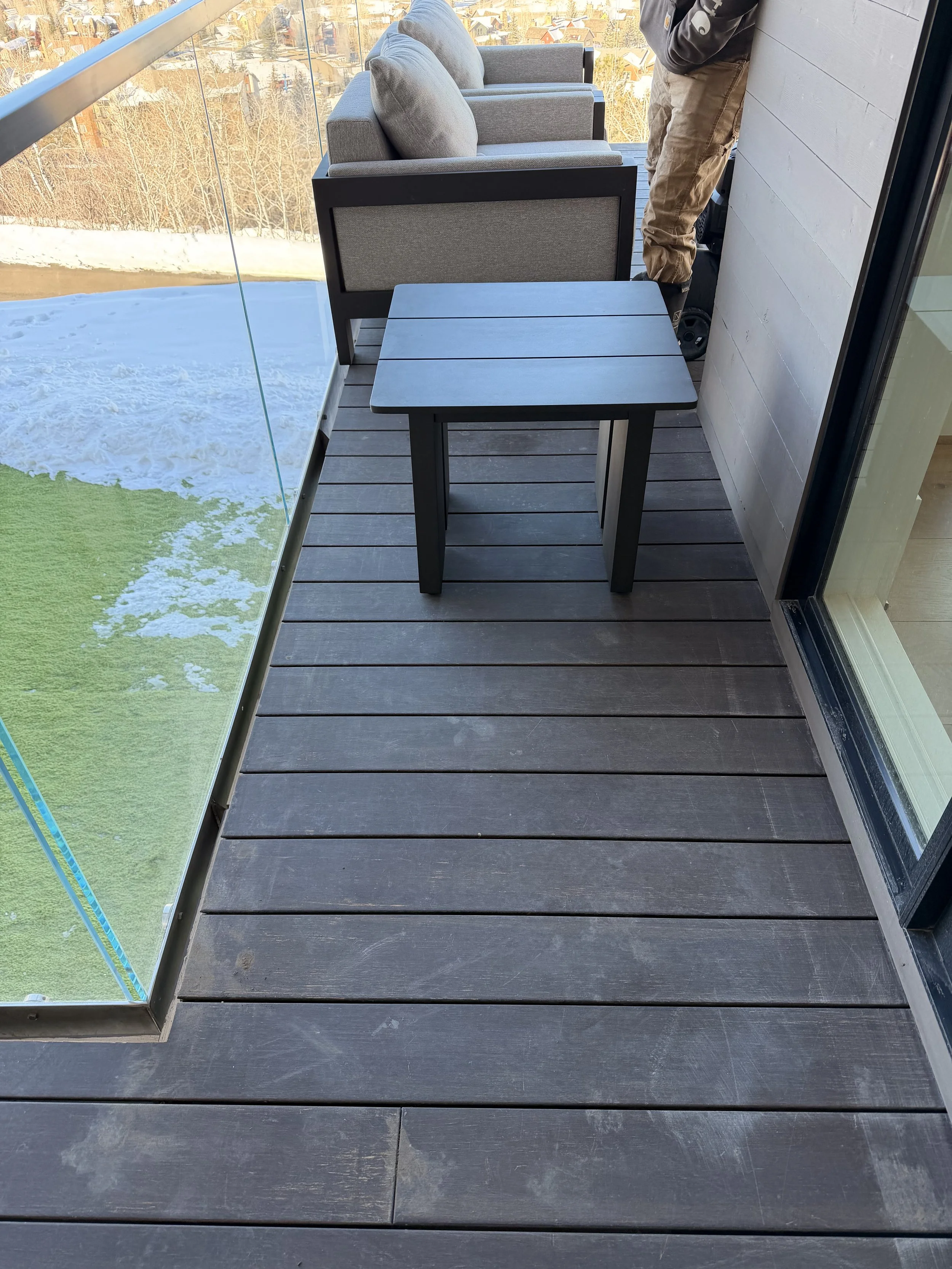 A balcony with dark wooden flooring, gray outdoor furniture including two armchairs and a small square table, and a glass railing revealing a snowy landscape and neighboring houses in the background.