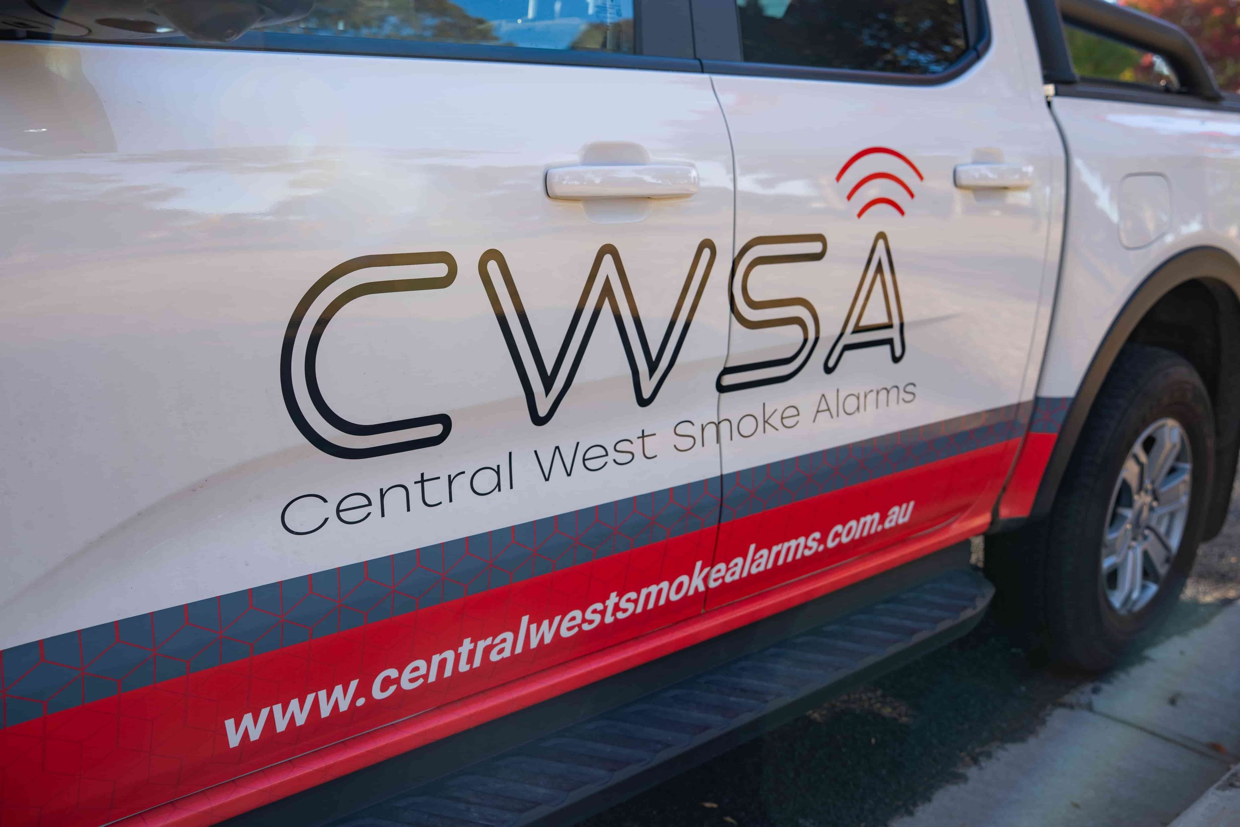 White vehicle with logo and text for Central West Smoke Alarms, including website URL, parked outdoors with trees reflected in the windows. Lithgow NSW