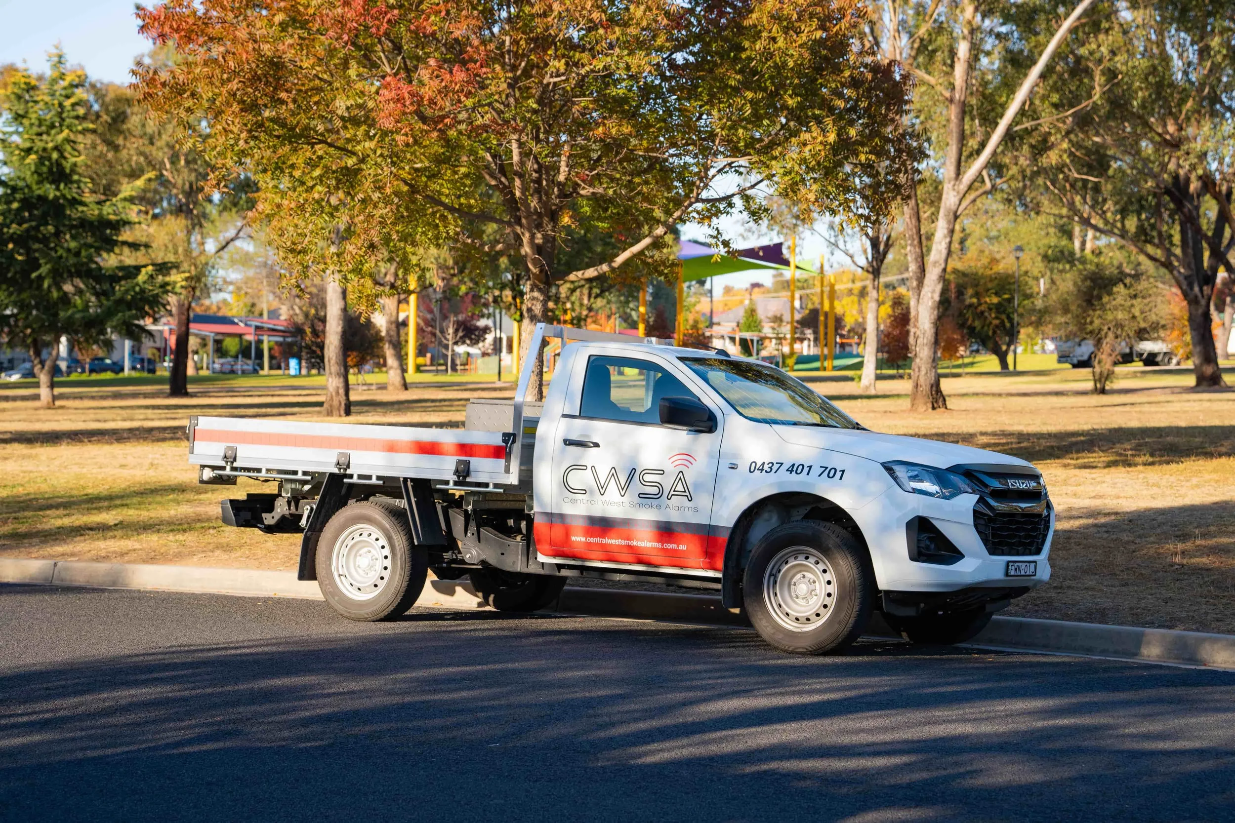 A white pickup truck with a logo reading 'CWSA Central West Smoke Alarms' parked on a street in a park with trees that have colorful fall leaves in Bathurst NSW