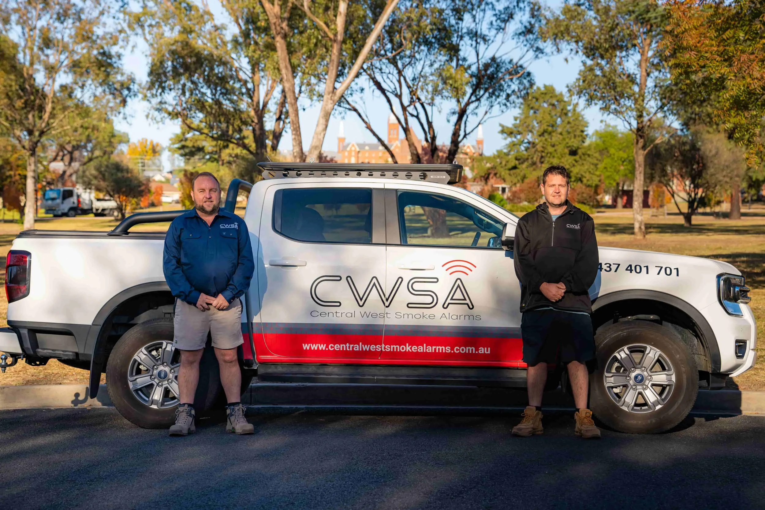 Two men standing in front of a white truck with CWSA logo and website, surrounded by trees and a building with towers in the background.