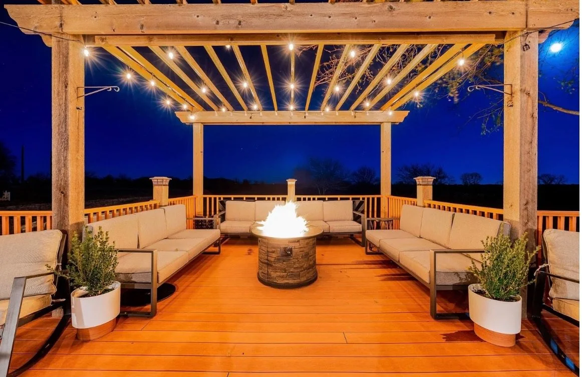 Outdoor deck at night with seating, fire pit, string lights, and a pergola overhead.