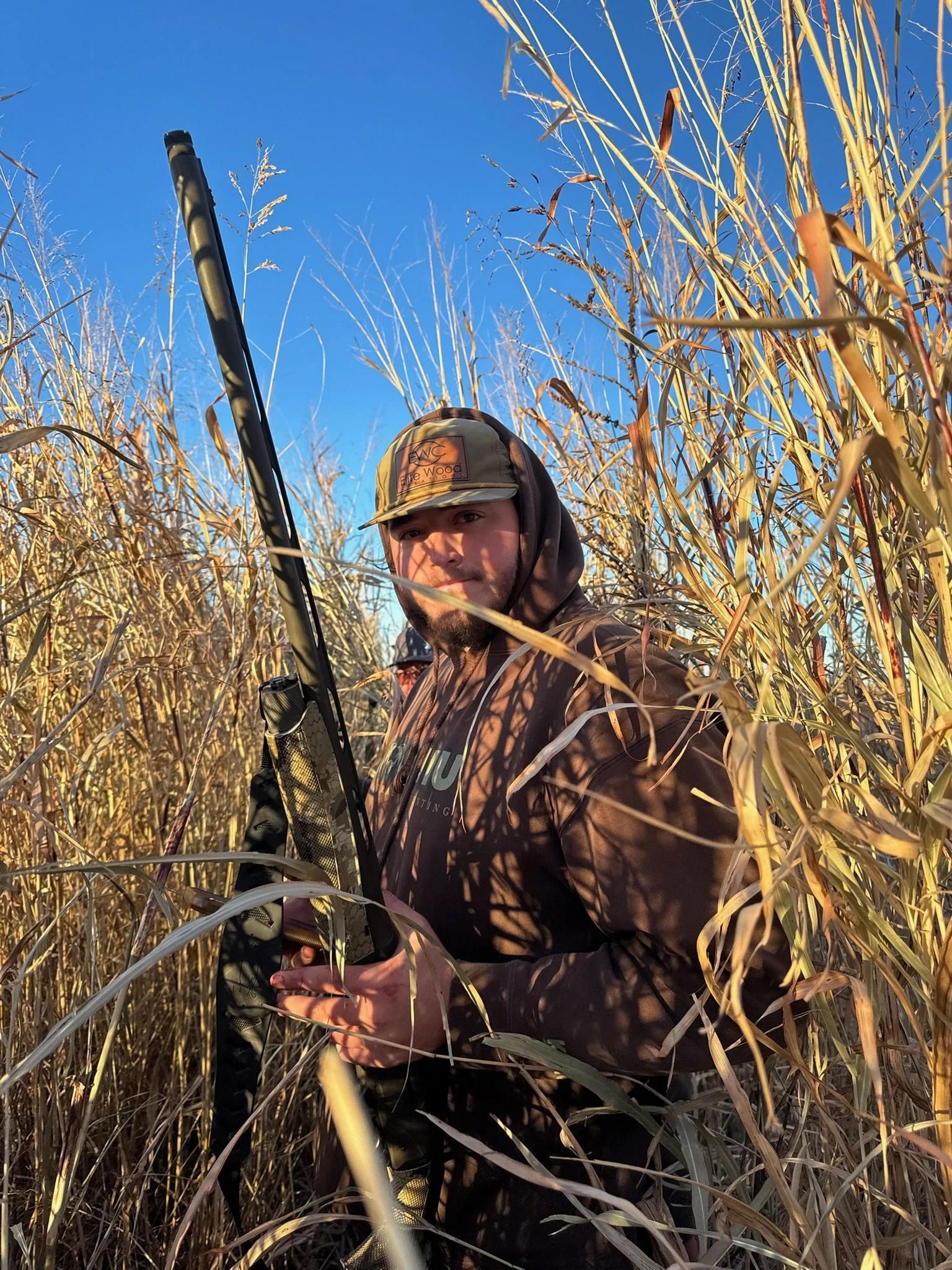 A man in camouflage clothing and a cap holding a hunting gun standing amidst tall, dry grass under a clear blue sky.