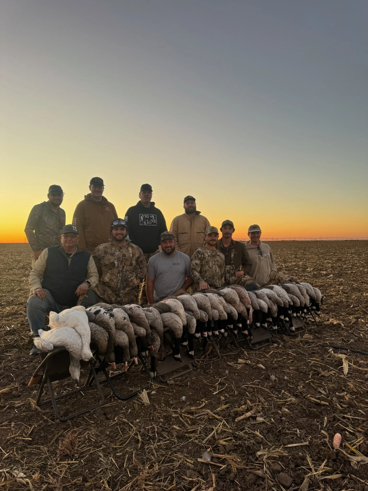 A group of ten hunters posing for a photo in a field at sunset, with a row of dead ducks and a white goose in front of them.