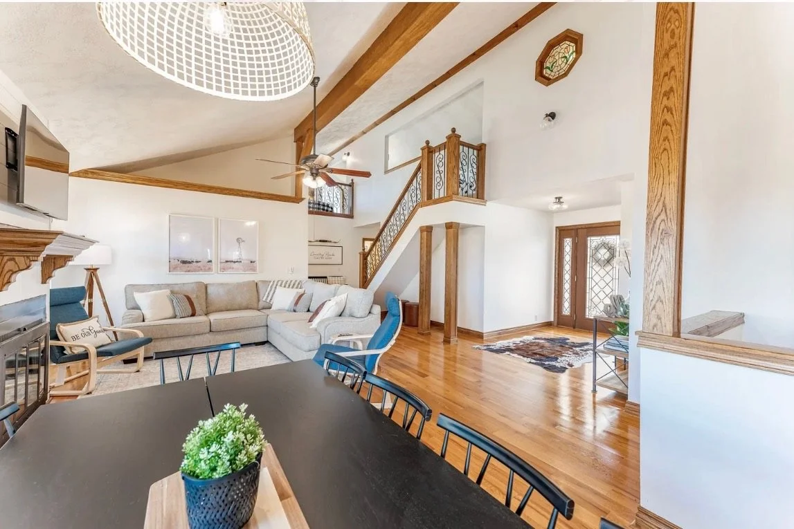 Living room with a beige sectional sofa, a blue armchair, and a dark wood dining table with chairs. Wooden accents and trim are visible on the ceiling, walls, and staircase. There are wall art, a ceiling fan, and a fireplace with a TV above it.
