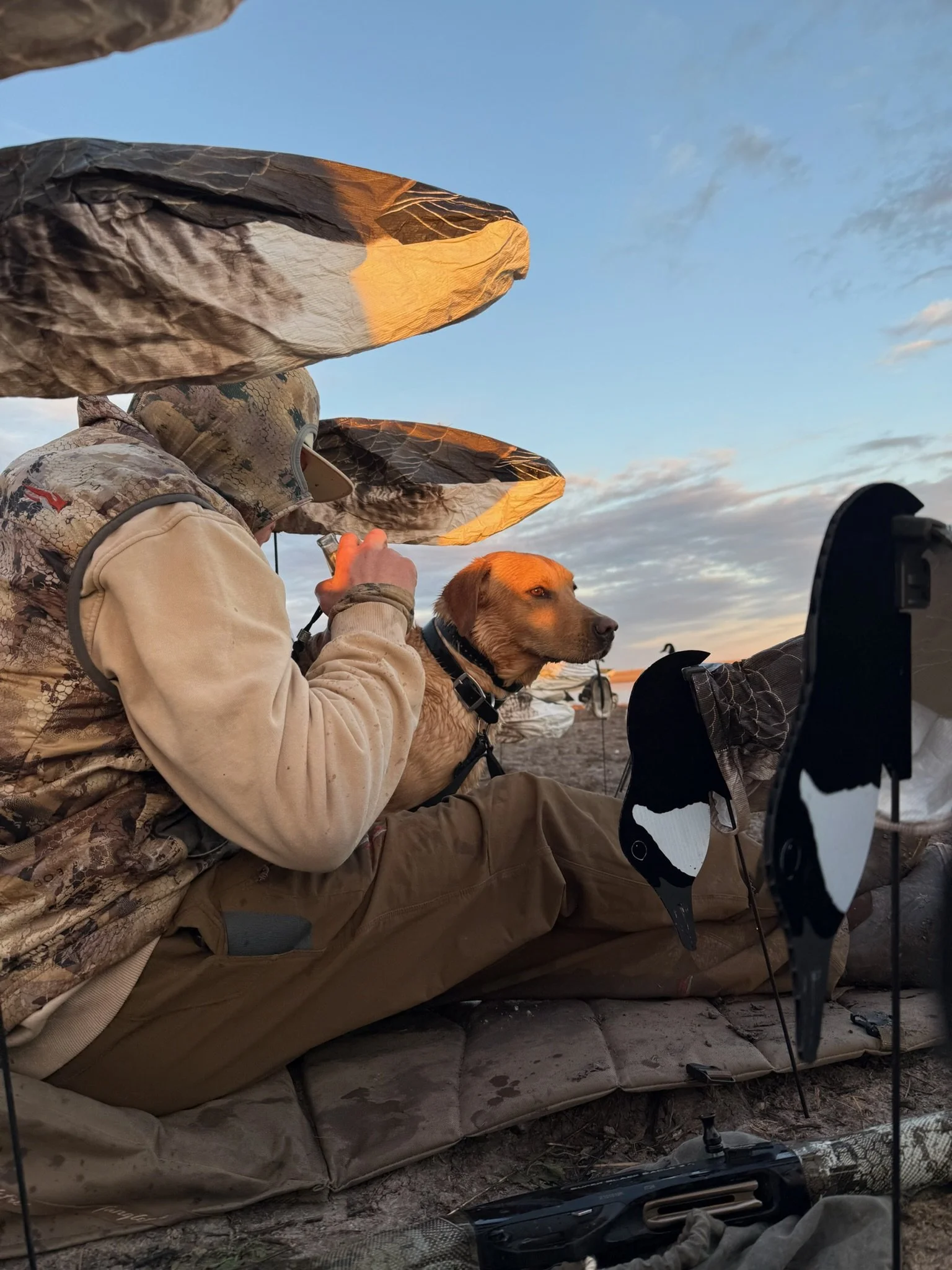 A person dressed in camouflage and beige clothing sitting on a mat outdoors at sunset with a dog, possibly a retriever, sitting next to them. The person is wearing a hat or camouflage head covering, and there are decoys around them, indicating a duck