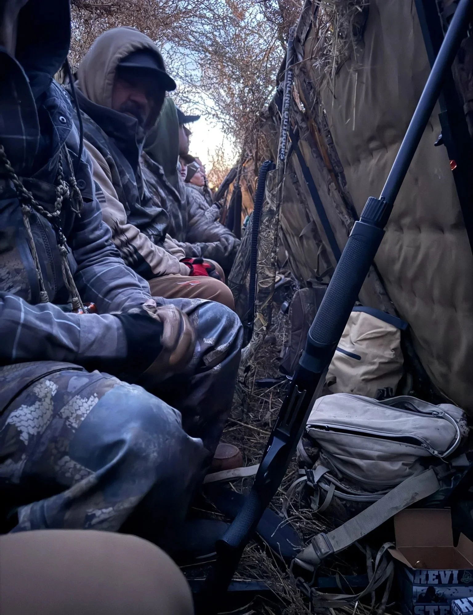 Group of people sitting outdoors in camouflage clothing, likely in a hunting blind, with equipment including a rifle and backpacks around them.