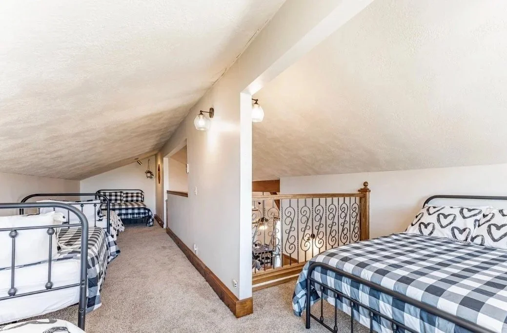 An attic bedroom with sloped ceiling, featuring multiple beds with black and white plaid bedding, a wooden railing overlooking the stairs, and wall-mounted lights.