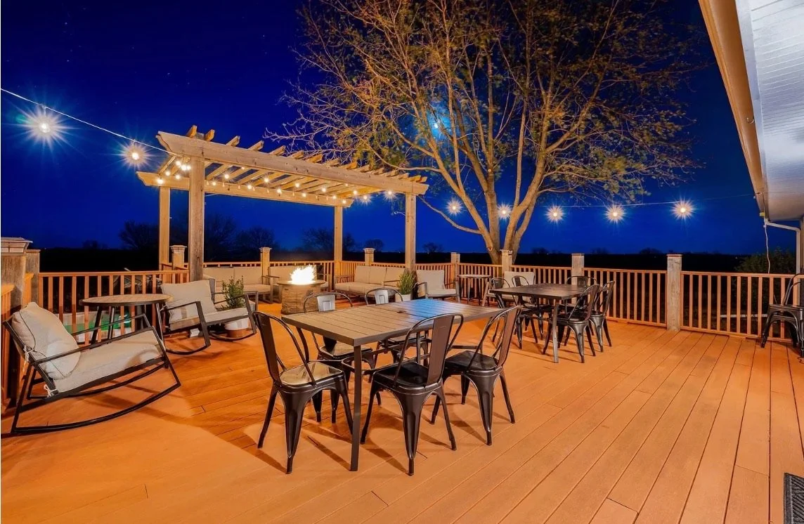 Outdoor deck with string lights, a tree, a fire pit, and sitting and dining furniture at night.