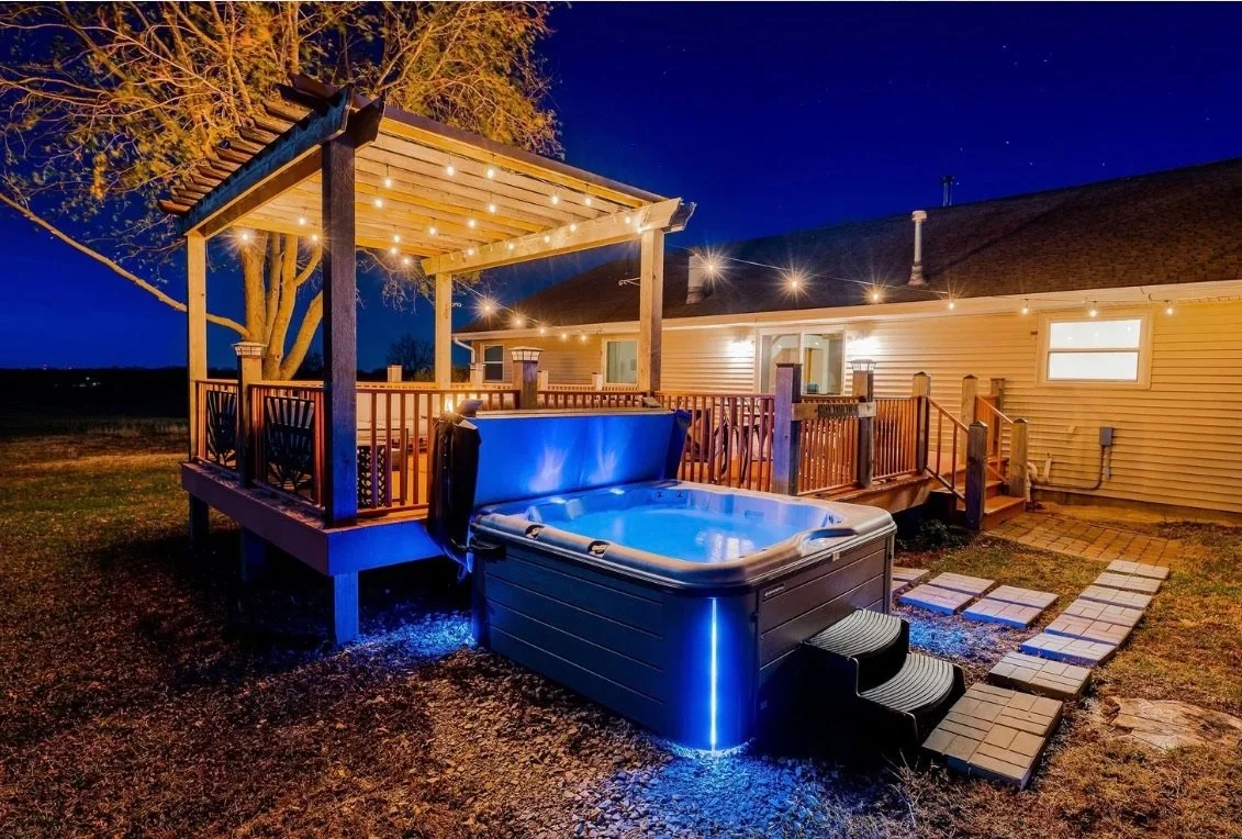 A backyard deck with string lights at night, featuring a hot tub with blue lighting, a stone pathway, and a house with exterior lights.