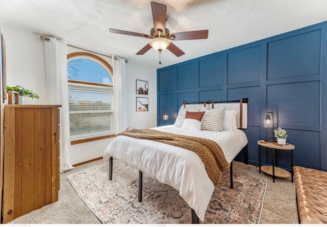 A bedroom with a white bed, a blue accent wall, wooden furniture, and a ceiling fan.