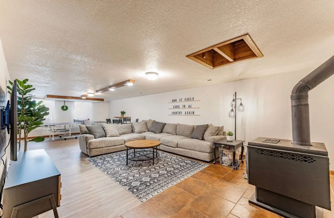 Living room with L-shaped sofa, wooden coffee table, patterned rug, wall sign, wood stove, potted plants, dining table with chairs, and ping pong table in the background