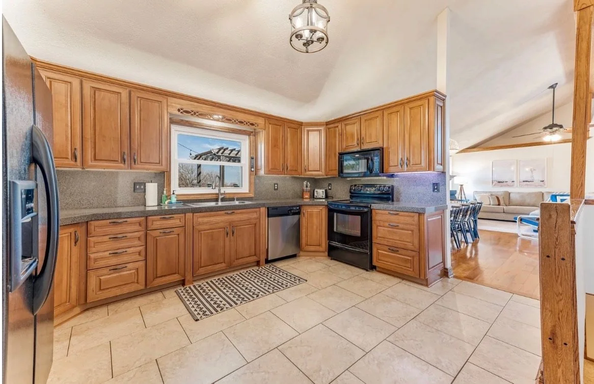 Kitchen with wooden cabinets, gray countertops, black stove, microwave, dishwasher, and a window above the sink showing an outdoor scene.