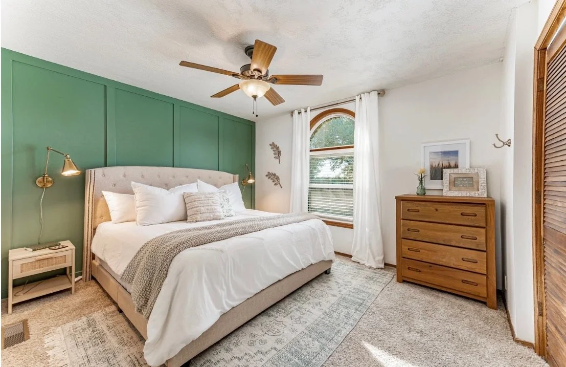 Bedroom with a beige bed, white bedding, cream-colored pillows, and a green accent wall. There are two wall-mounted lamps on either side of the bed, a bedside table on the left, a wood dresser on the right, and a window with white curtains and an arched top.