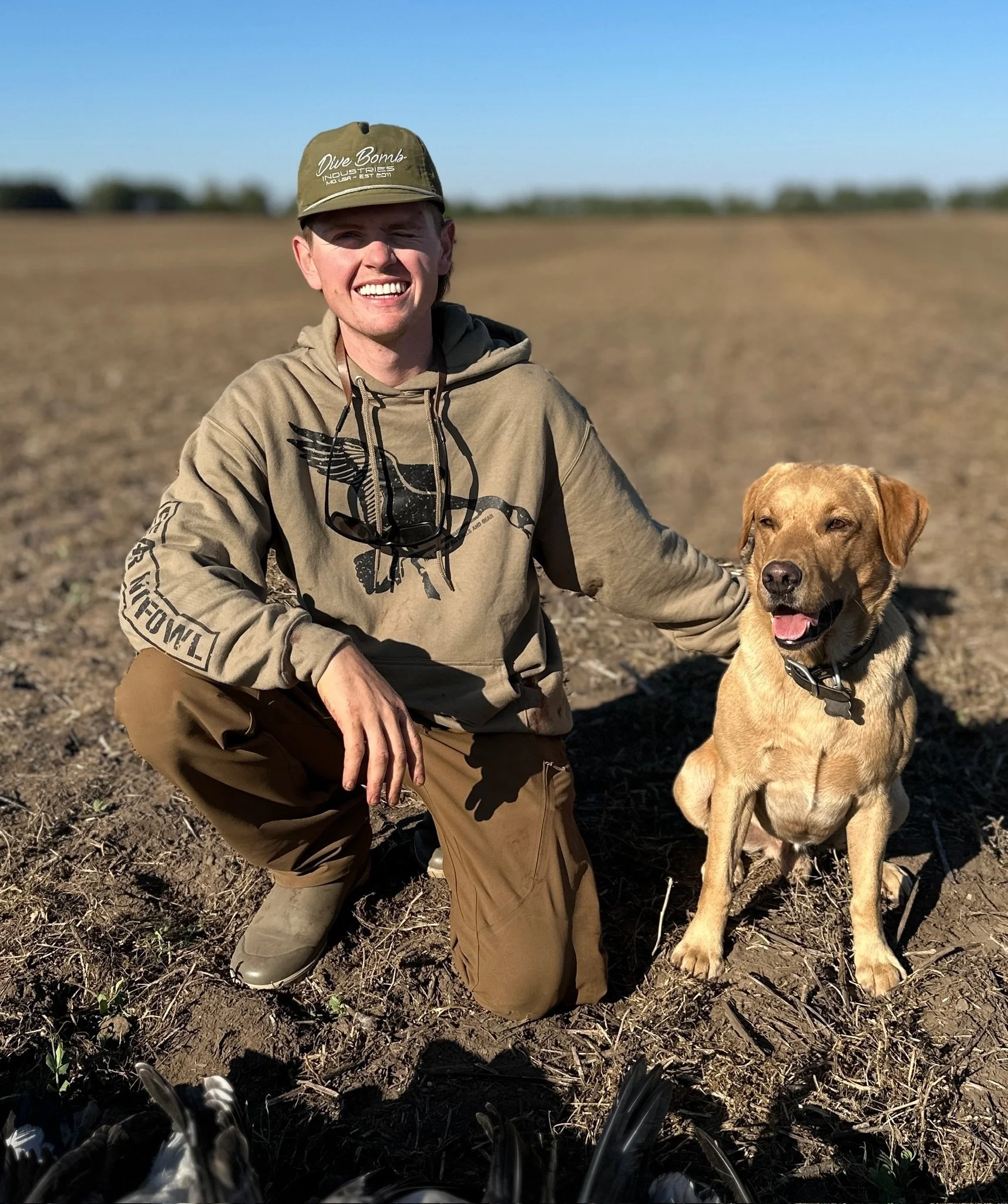 A young man kneeling in a field with his dog, both smiling at the camera on a sunny day.