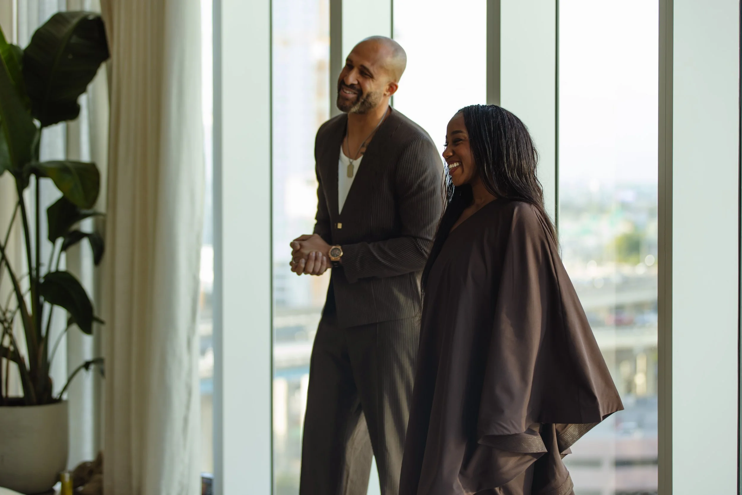 A man and a woman standing indoors in front of large windows, smiling and engaged in conversation.