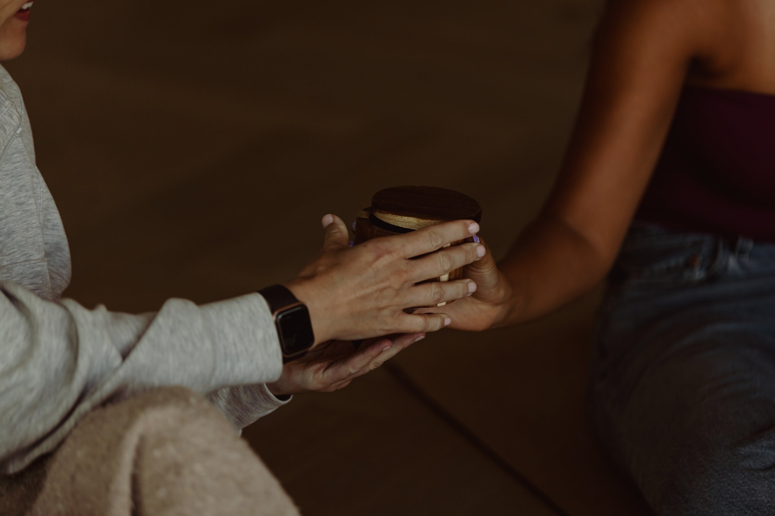 Two people exchange an item, one person with light skin wearing a gray long sleeve shirt and a black smartwatch, the other with dark skin wearing a maroon top, denim jeans, and a black bracelet, in an indoor setting.