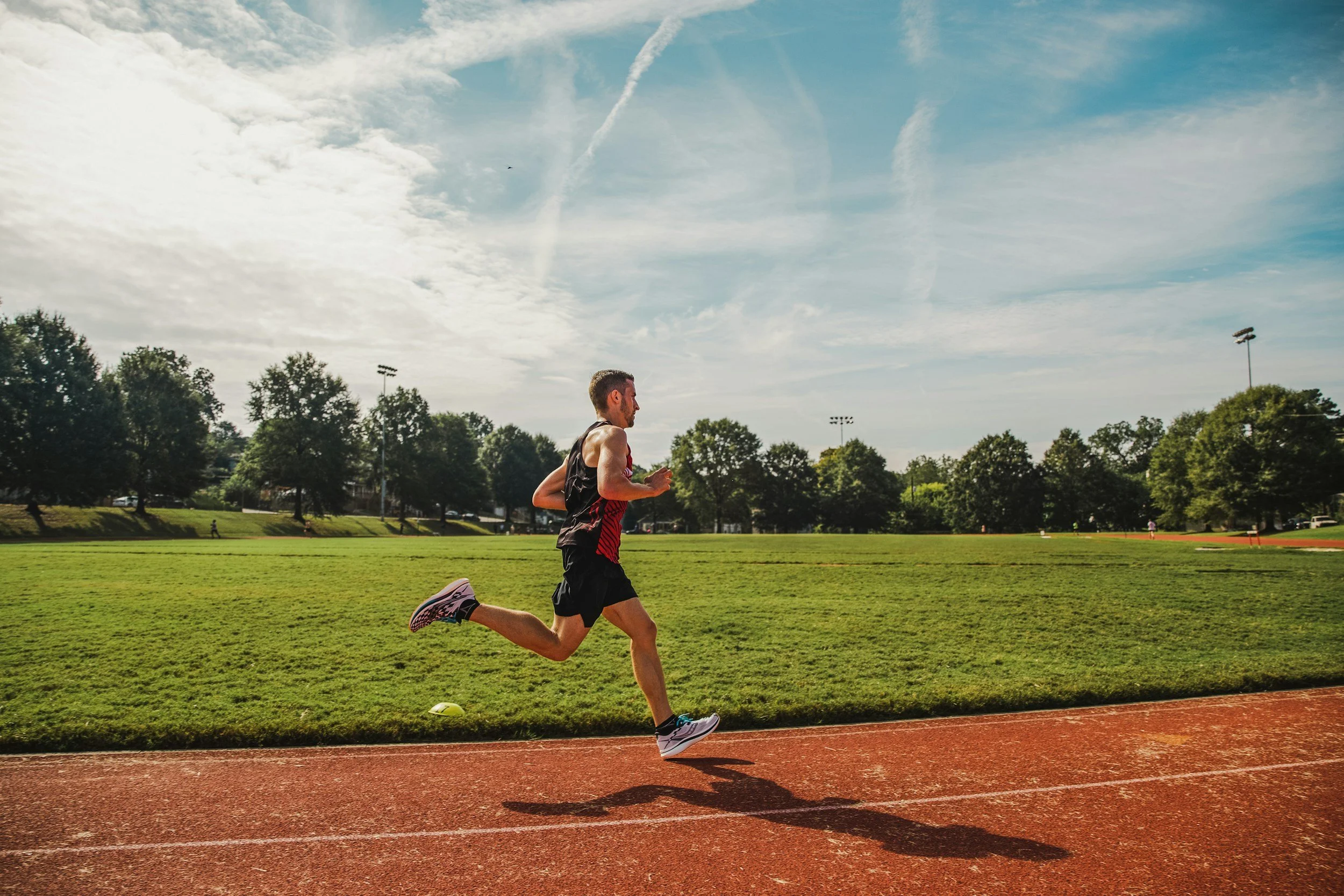 A man running on an outdoor track in a park on a sunny day with a partly cloudy sky and trees in the background.