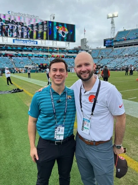 Two men smiling and standing on a football field at EverBank Stadium, with large screens and stadium seating in the background, in cloudy weather.