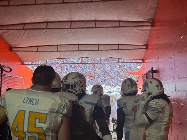 Football players walking onto the field through a tunnel with a crowd in the background.