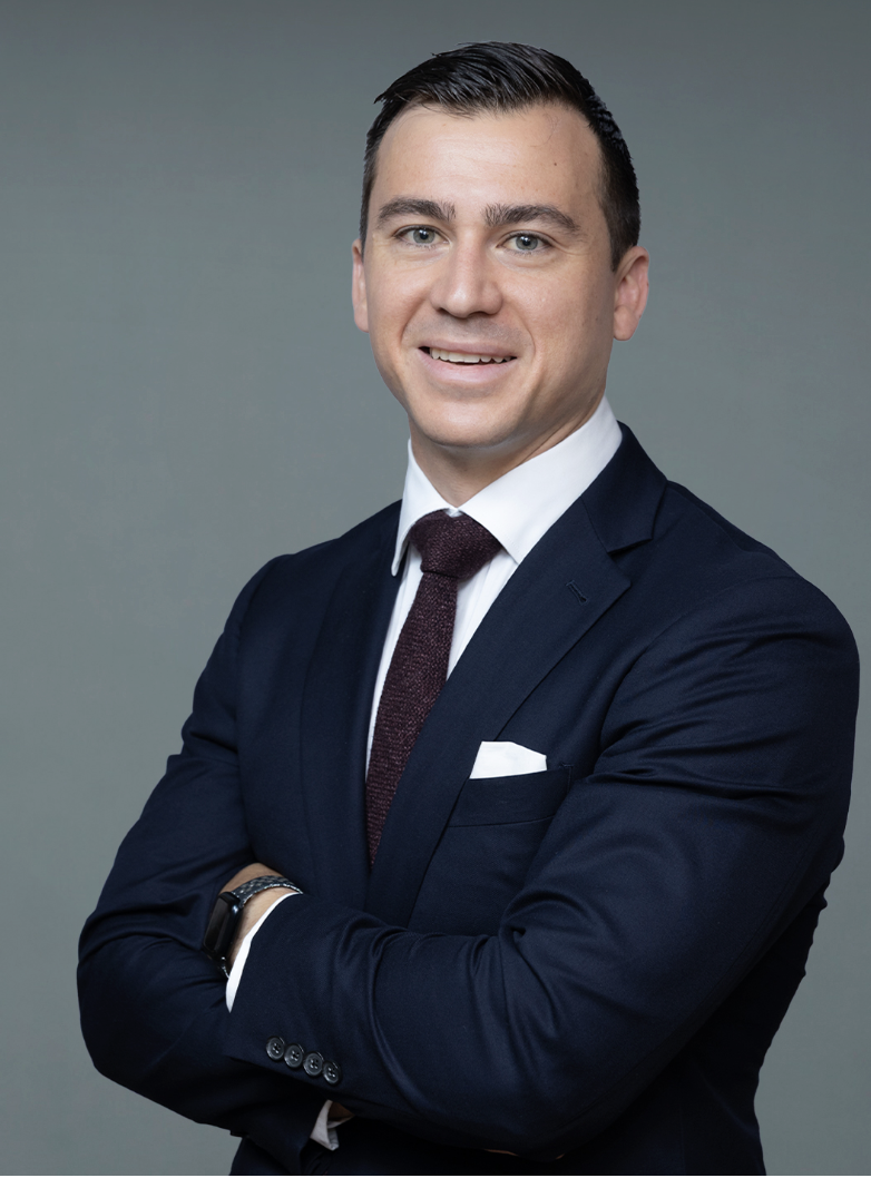 A professional man in a dark blue suit, white dress shirt, and maroon tie standing with arms crossed against a gray background.